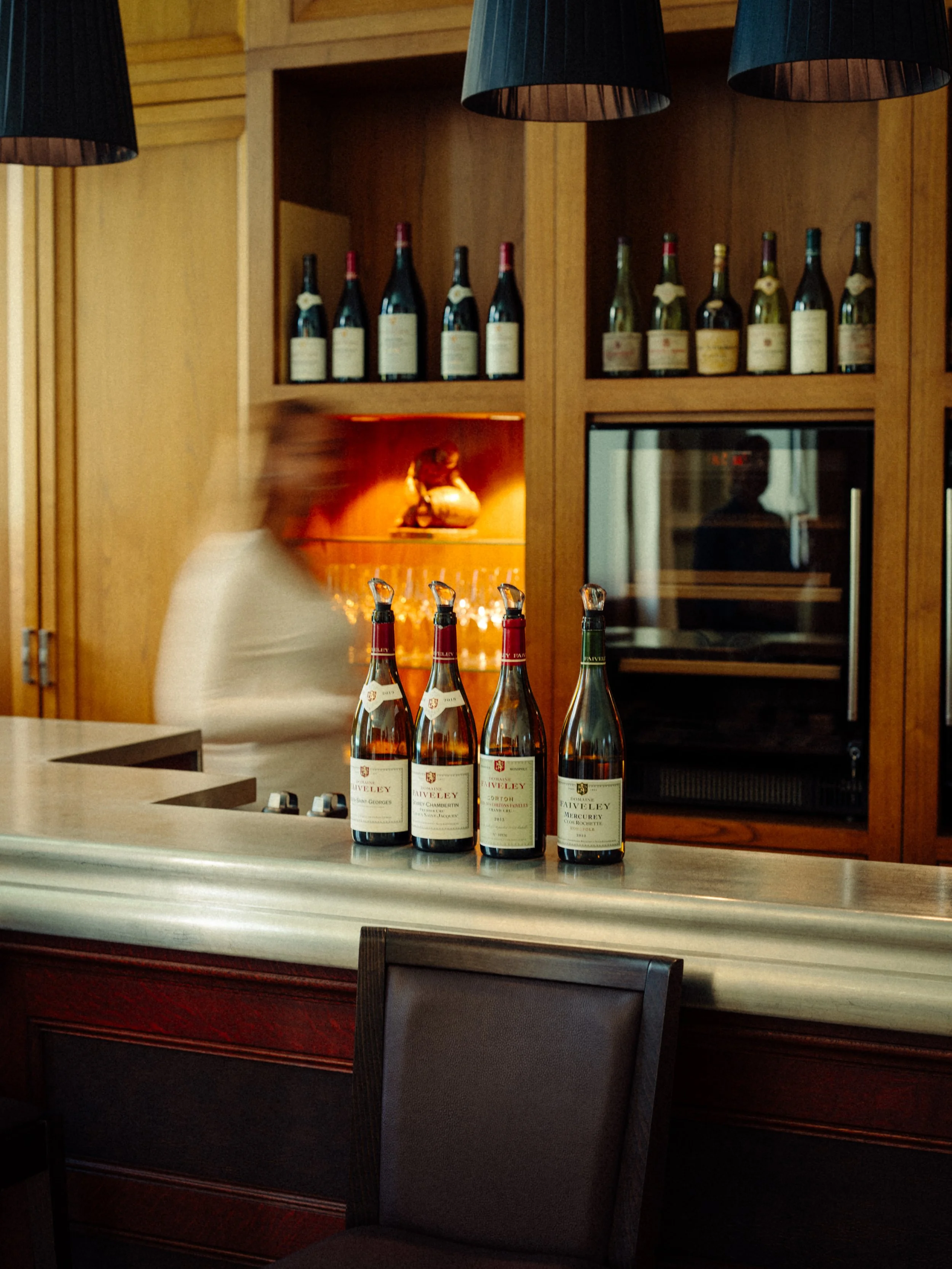 Bar avec bouteilles de vin en arrière-plan, un homme flou derrière le comptoir, bouteilles de vin sur le comptoir, éclairage chaud, ambiance chaleureuse.