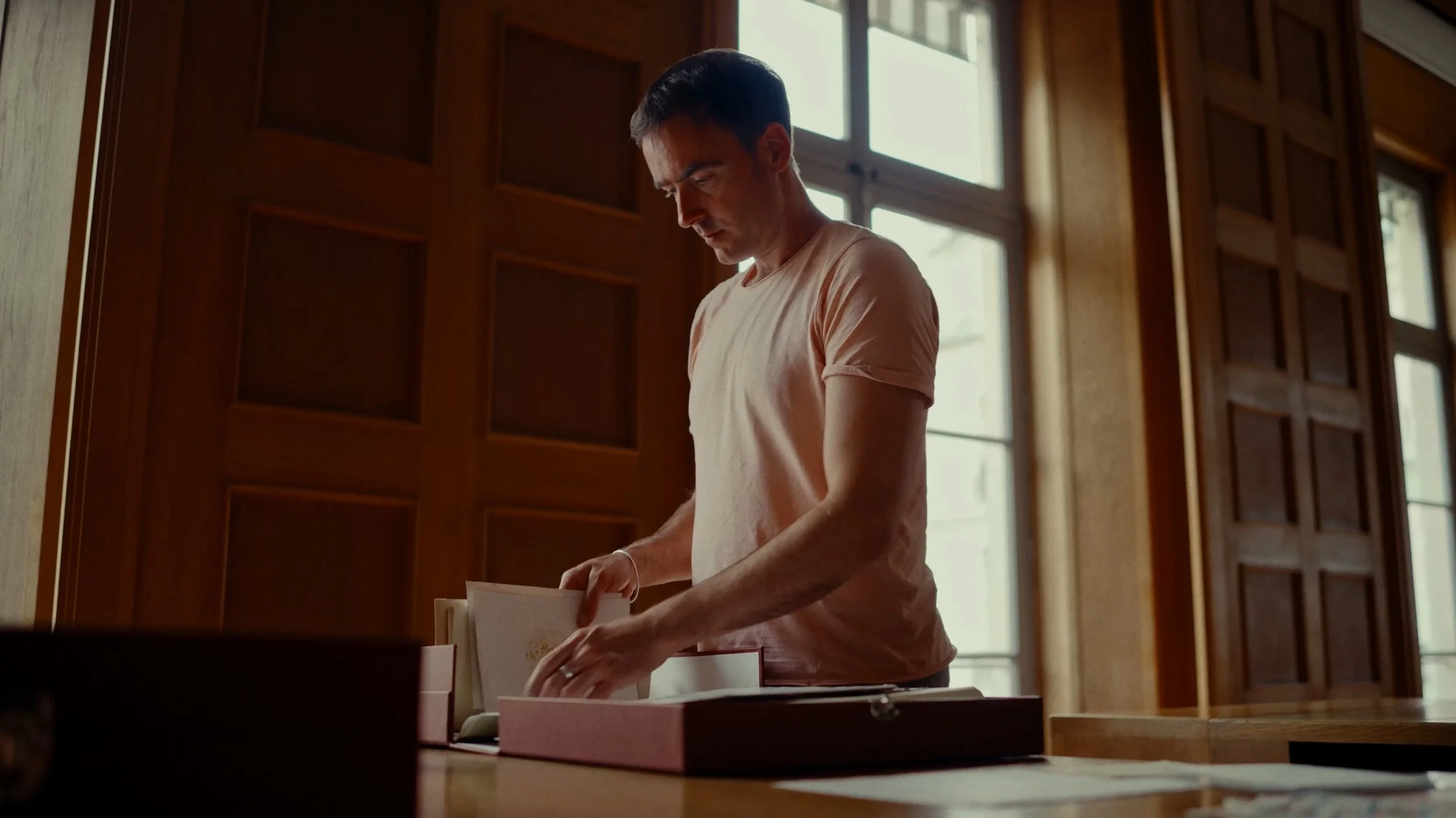 Un homme regarde à travers un classeur ouvert sur une table en bois dans une pièce avec de grandes fenêtres laissant entrer la lumière naturelle, murs en bois.