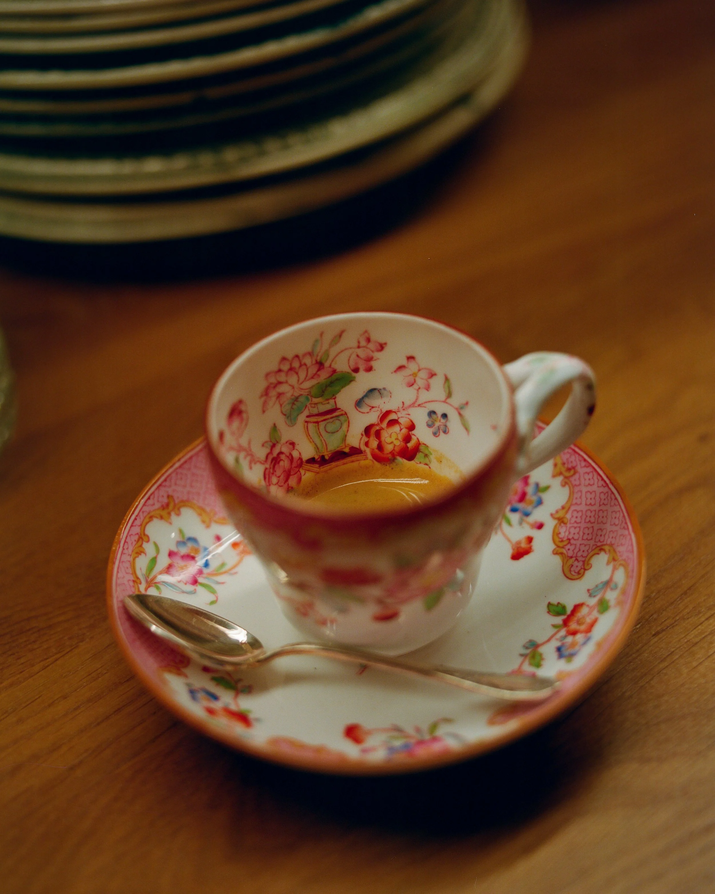 Une tasse en porcelaine avec un motif floral coloré, accompagnée d'une petite cuillère, repose sur une soucoupe assortie, placée sur une table en bois.