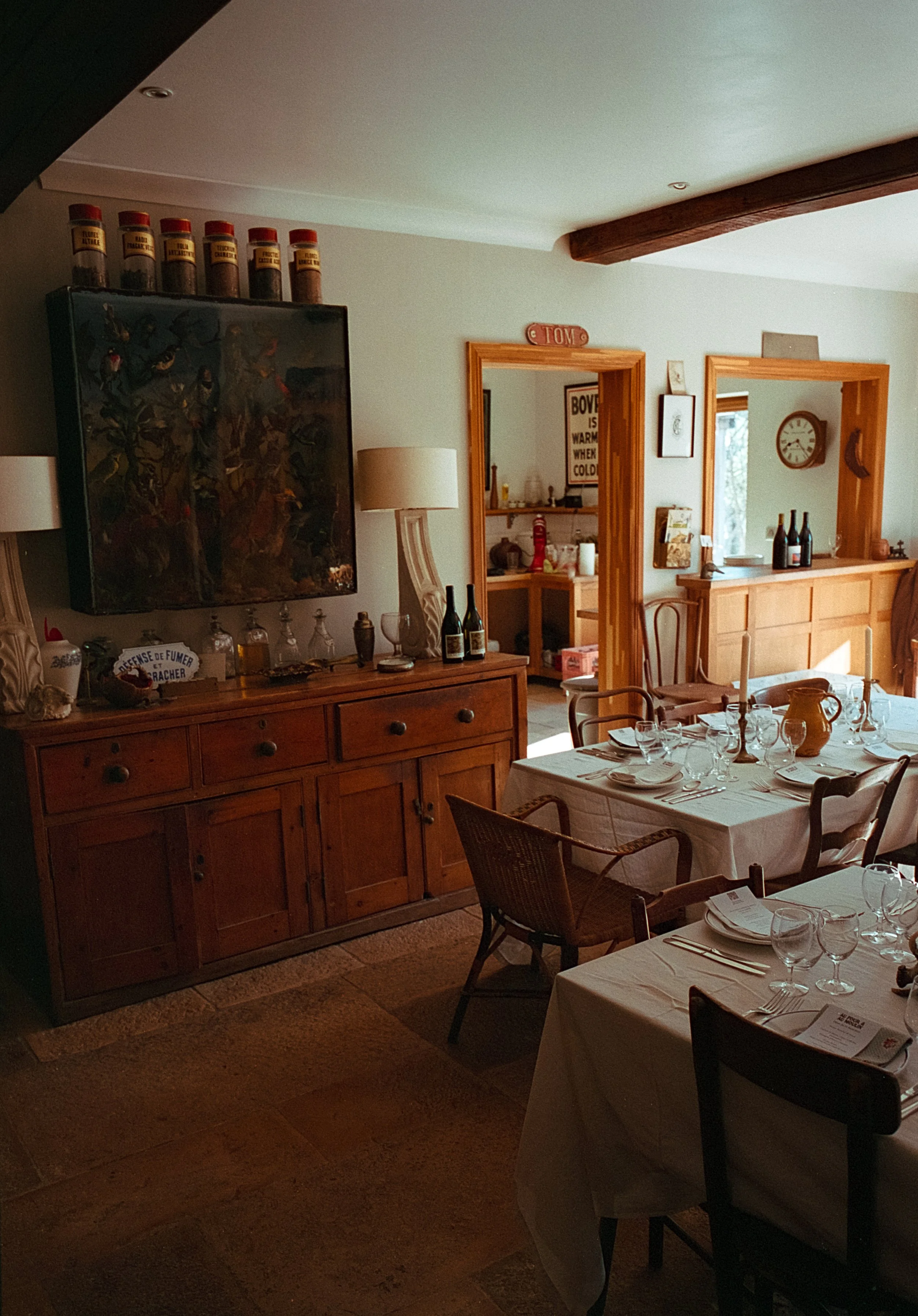 Salle à manger décorée avec une grande table et des chaises, un buffet en bois, des lampes, des bouteilles de vin, et des éléments de décoration vintage.