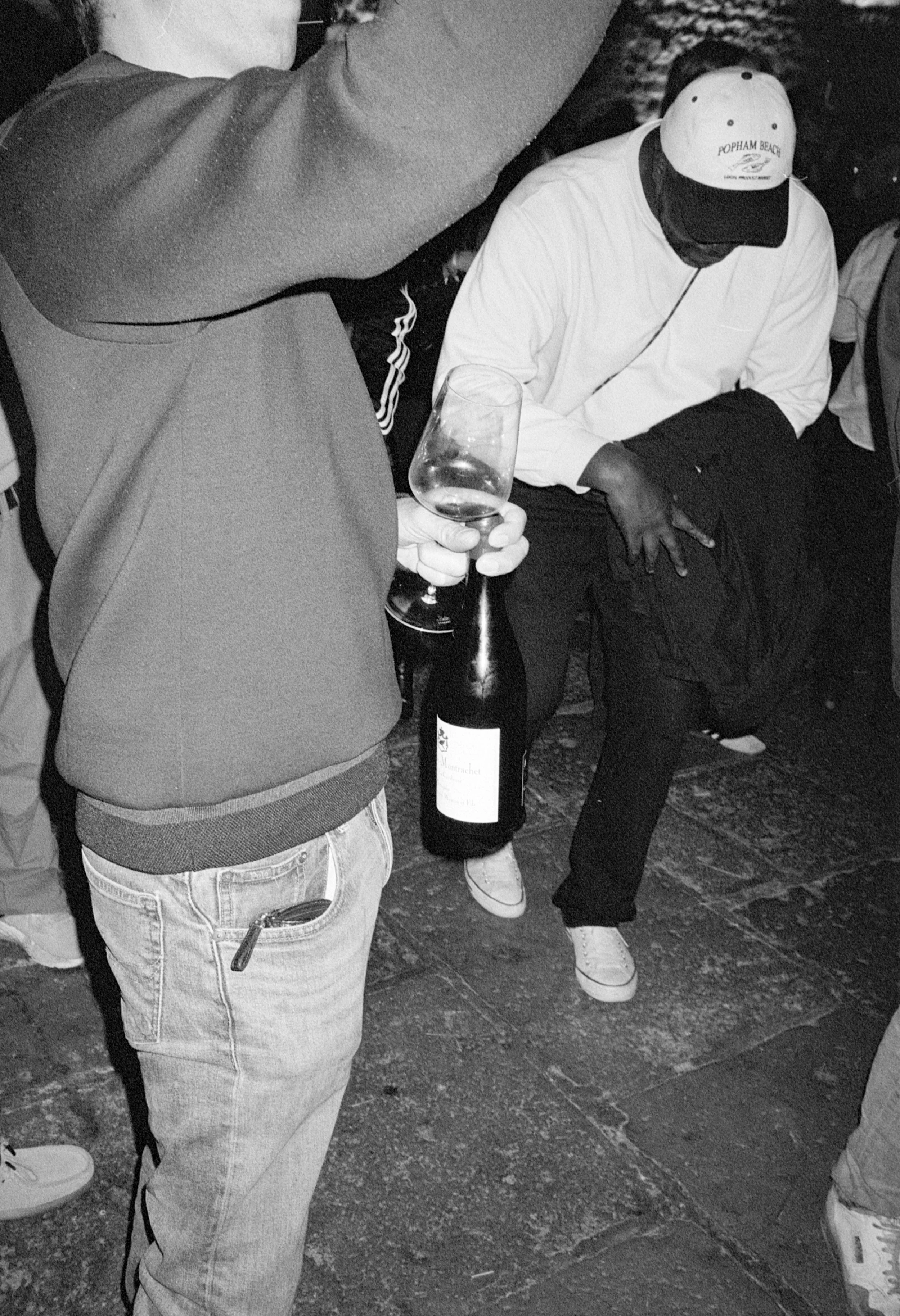 Deux personnes lors d'une rencontre sociale, une tenant un verre de vin et une autre assise, portant une casquette avec l'inscription 'Popham Beach'.