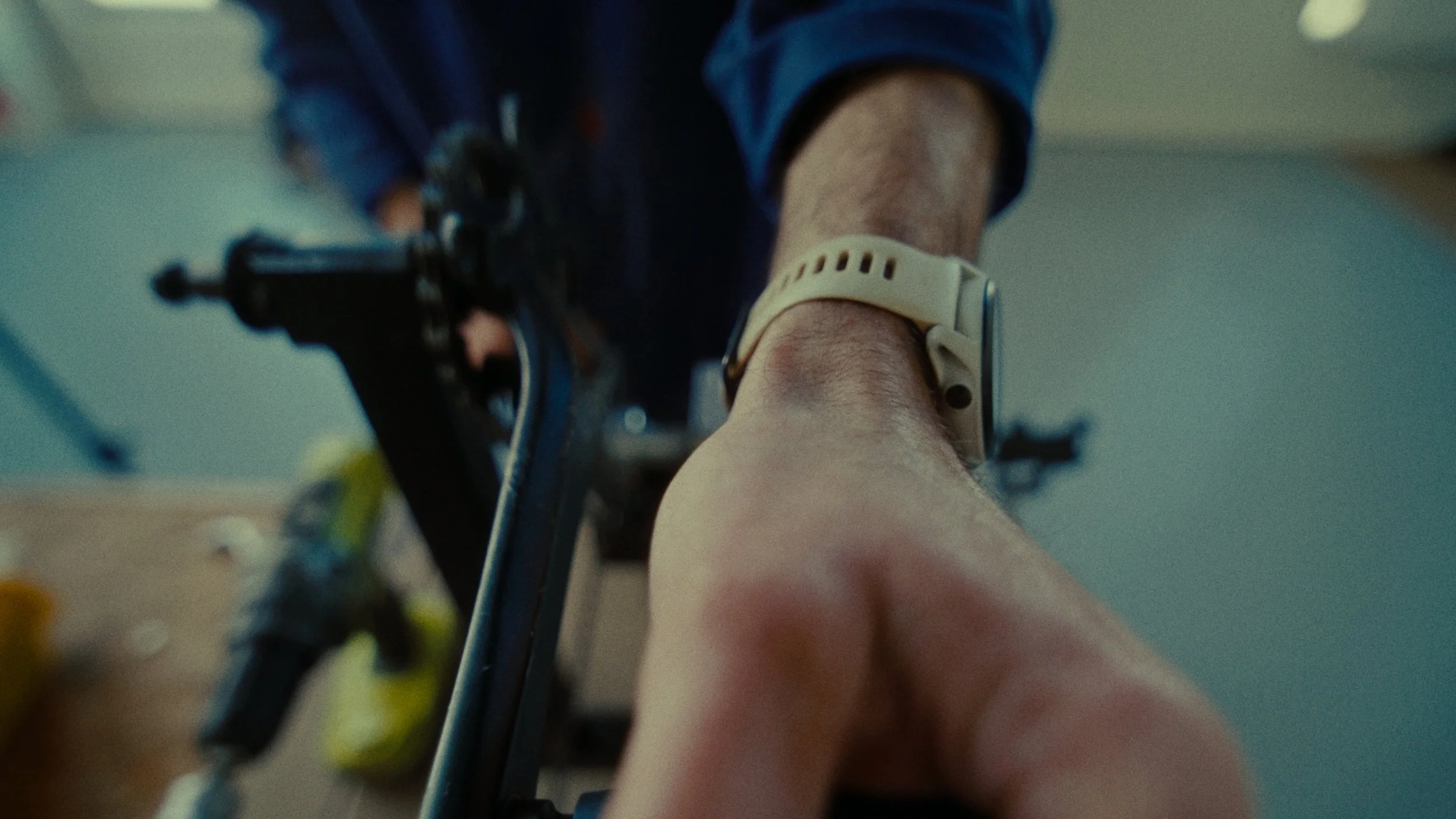 Prise de vue en contre-plongée montrant un bras d'adulte portant une montre ou un bracelet en plastique de couleur beige, avec un vélo devant lui, dans un intérieur. La photo est floue avec un fond neutre.