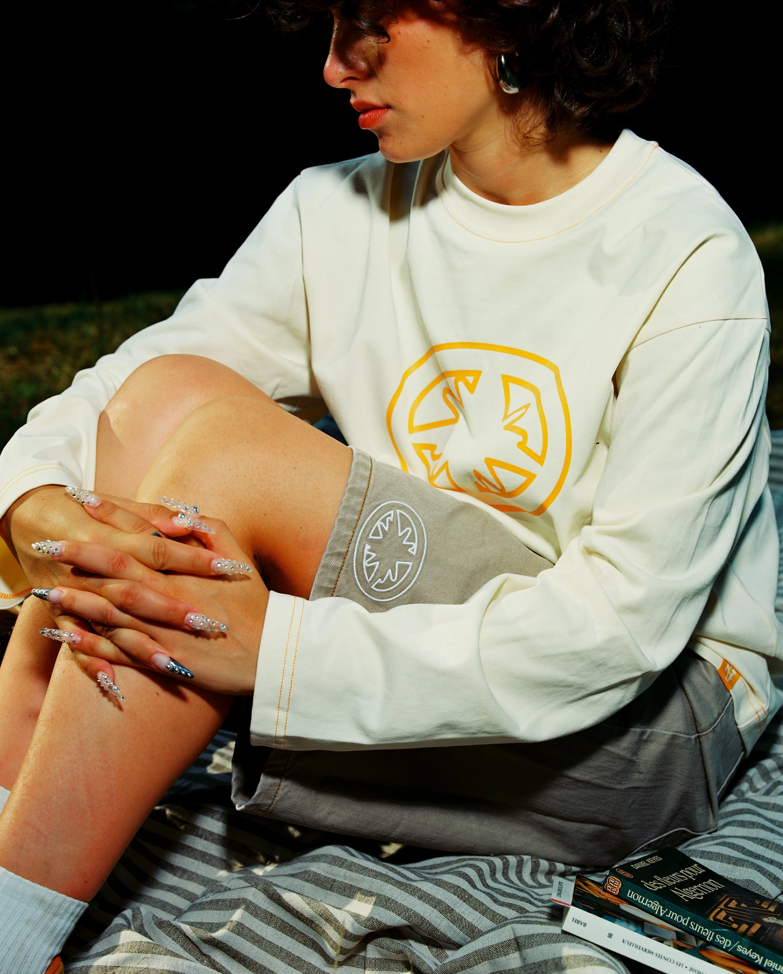 Une jeune femme assise sur une couverture, portant un sweat à capuche beige clair avec un logo orange et un short gris clair avec un logo blanc. Elle tient une jambe avec ses mains, portant des ongles décorés de strass. Des livres sont posés sur la c