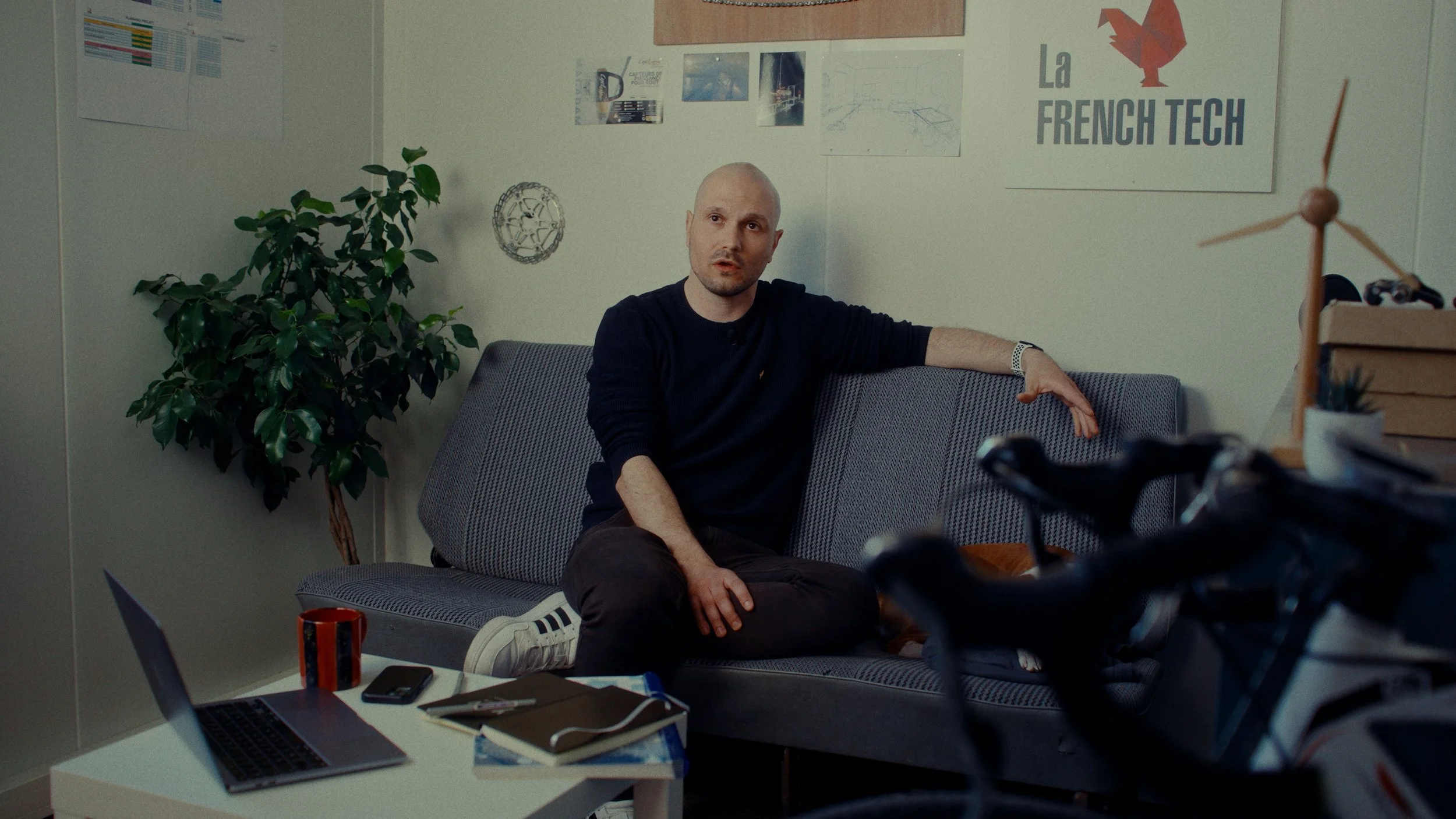 Un homme assis sur un canapé dans un bureau, entouré de plantes, de posters et de brainstorming. Des objets de bureau, un ordinateur portable, un carnet, une tasse et un téléphone sont visibles sur une table devant lui.