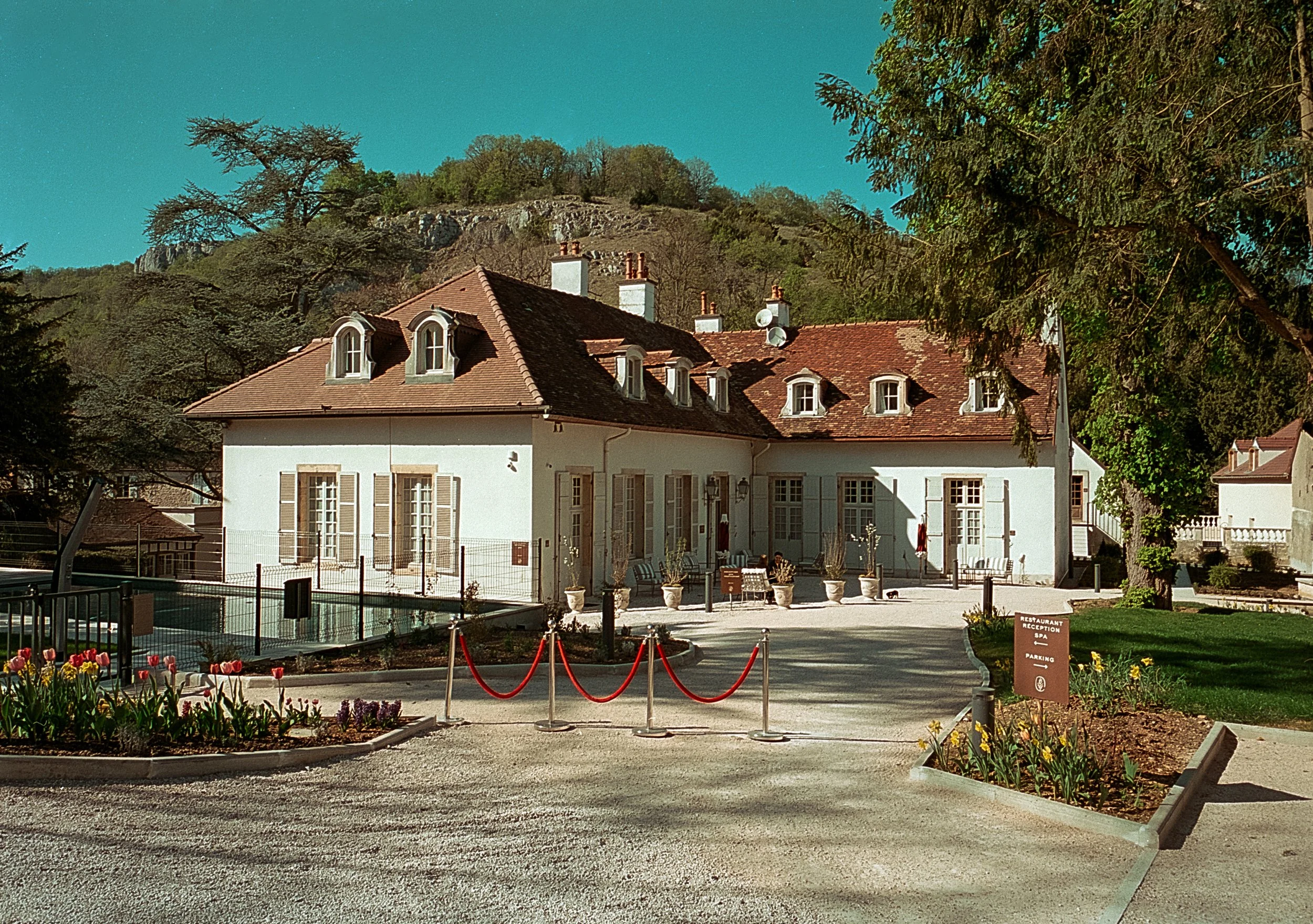 Maison blanche à toit rouge dans un jardin avec arbres, fleurs et rosiers, entourée d'une clôture et située au pied d'une colline avec des arbres.