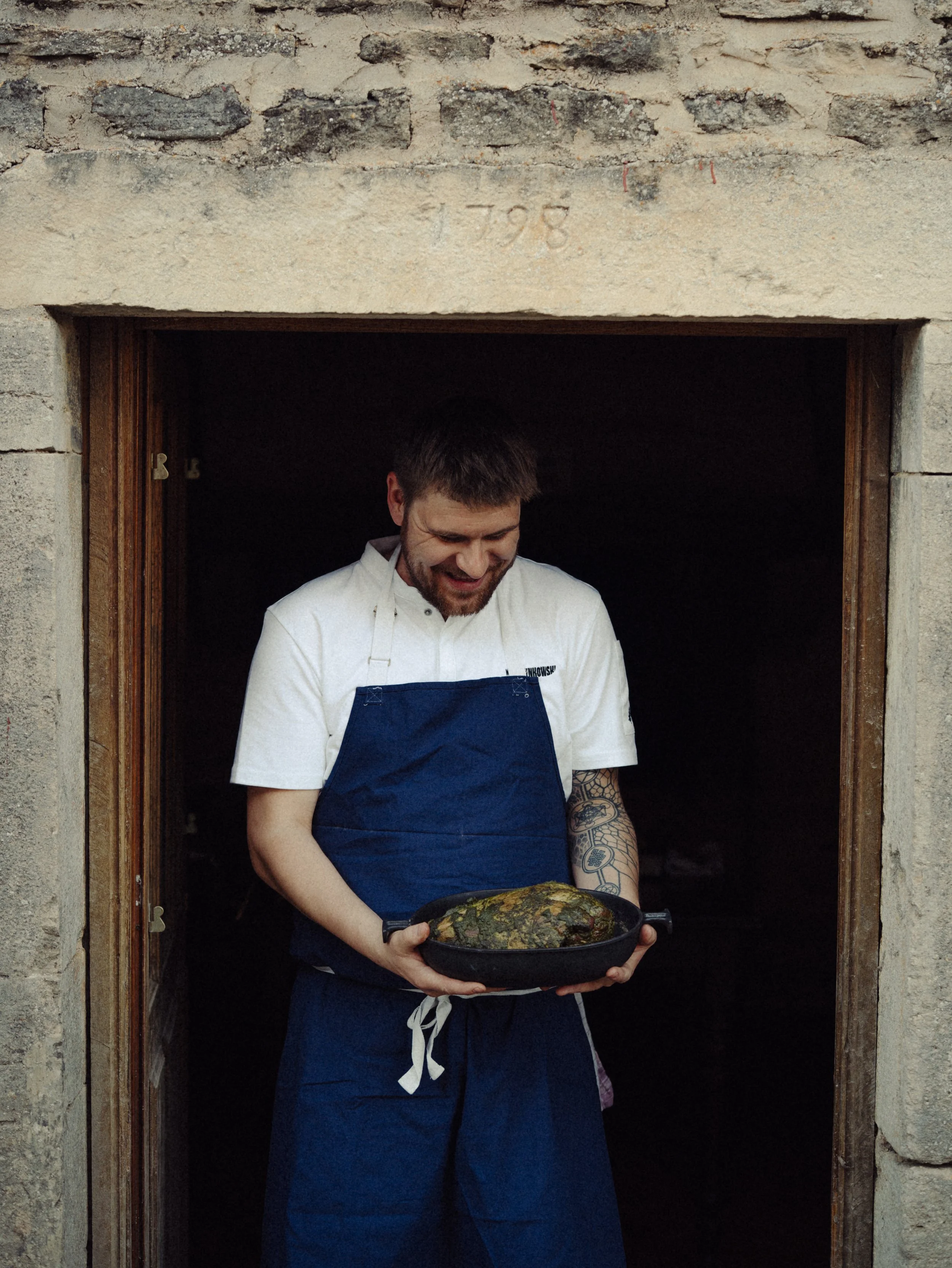 Un chef souriant tenant un plat avec une pièce de viande ou de poisson cuite, dans une cuisine.