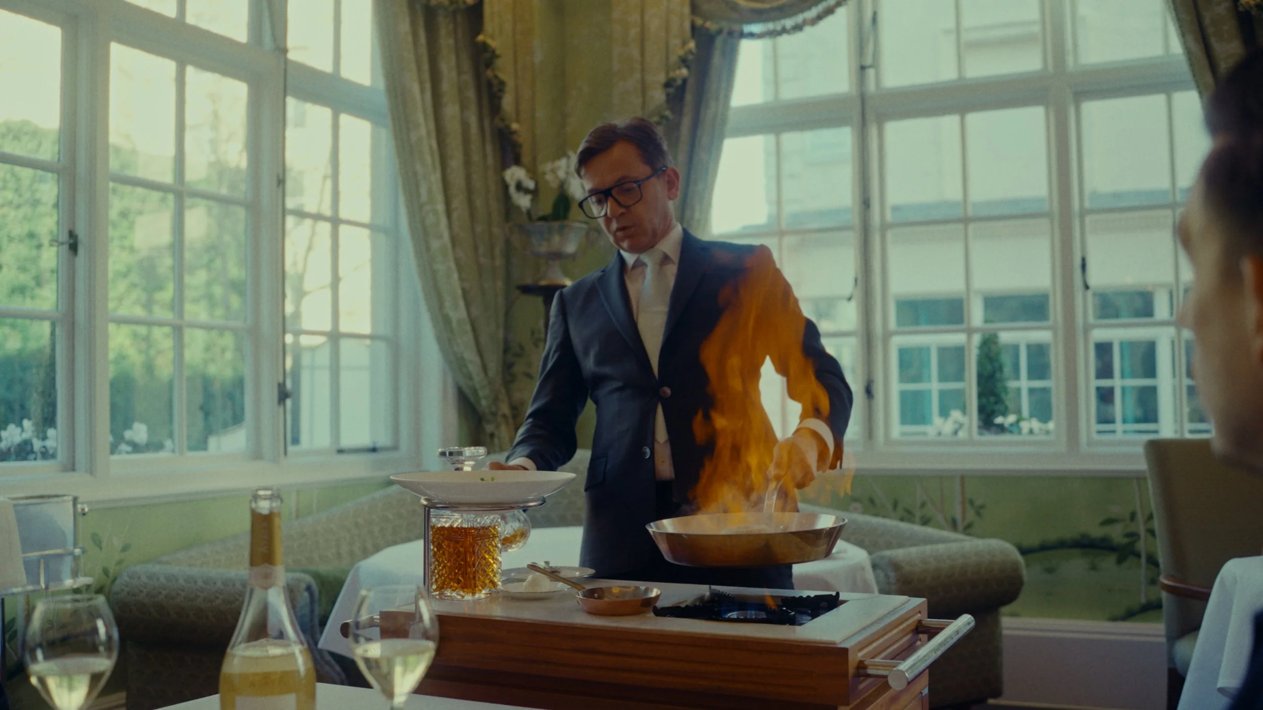 Un homme en costume prépare un plat en le flambant dans une cuisine de restaurant, avec des fenêtres grandes laissant entrer la lumière naturelle.