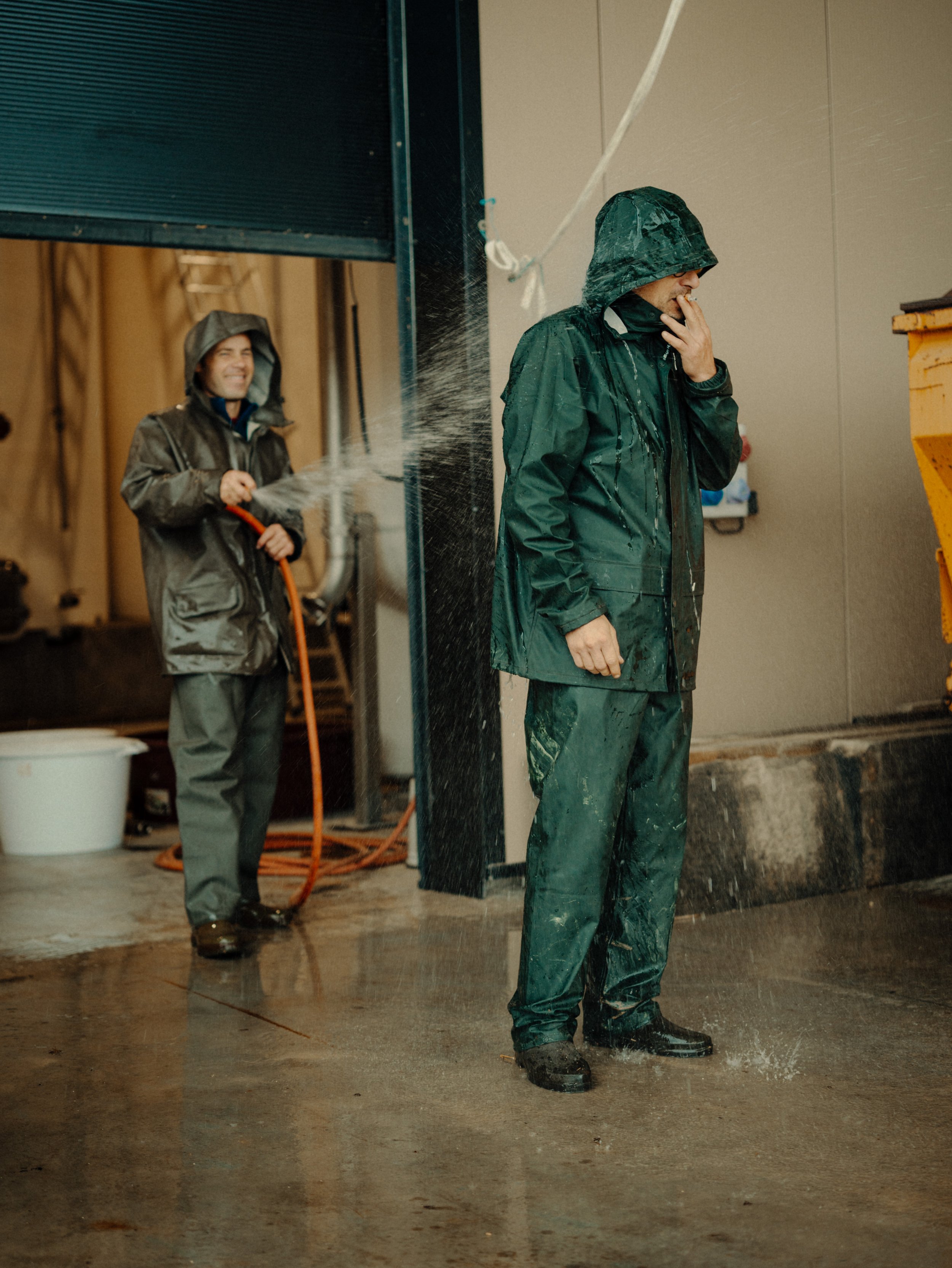 Deux hommes en vêtements de pluie noirs, un avec une capuche, se lavant sous une forte pluie artificielle à l'extérieur d'un bâtiment.
