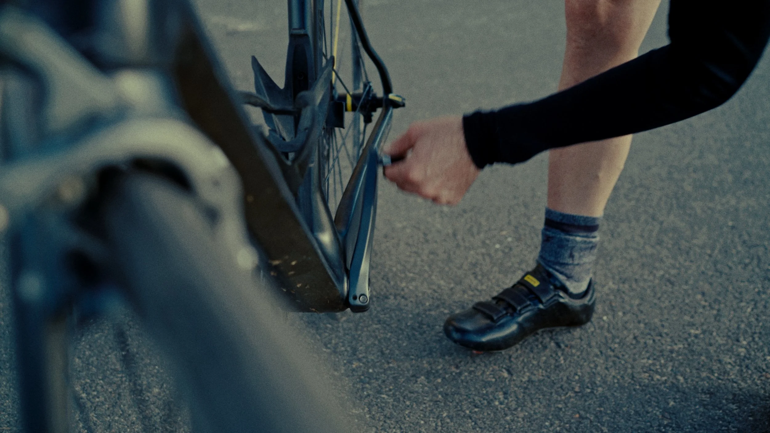 Personne ajustant la transmission d'un vélo, portant des chaussures de cyclisme, sur une surface asphaltée.