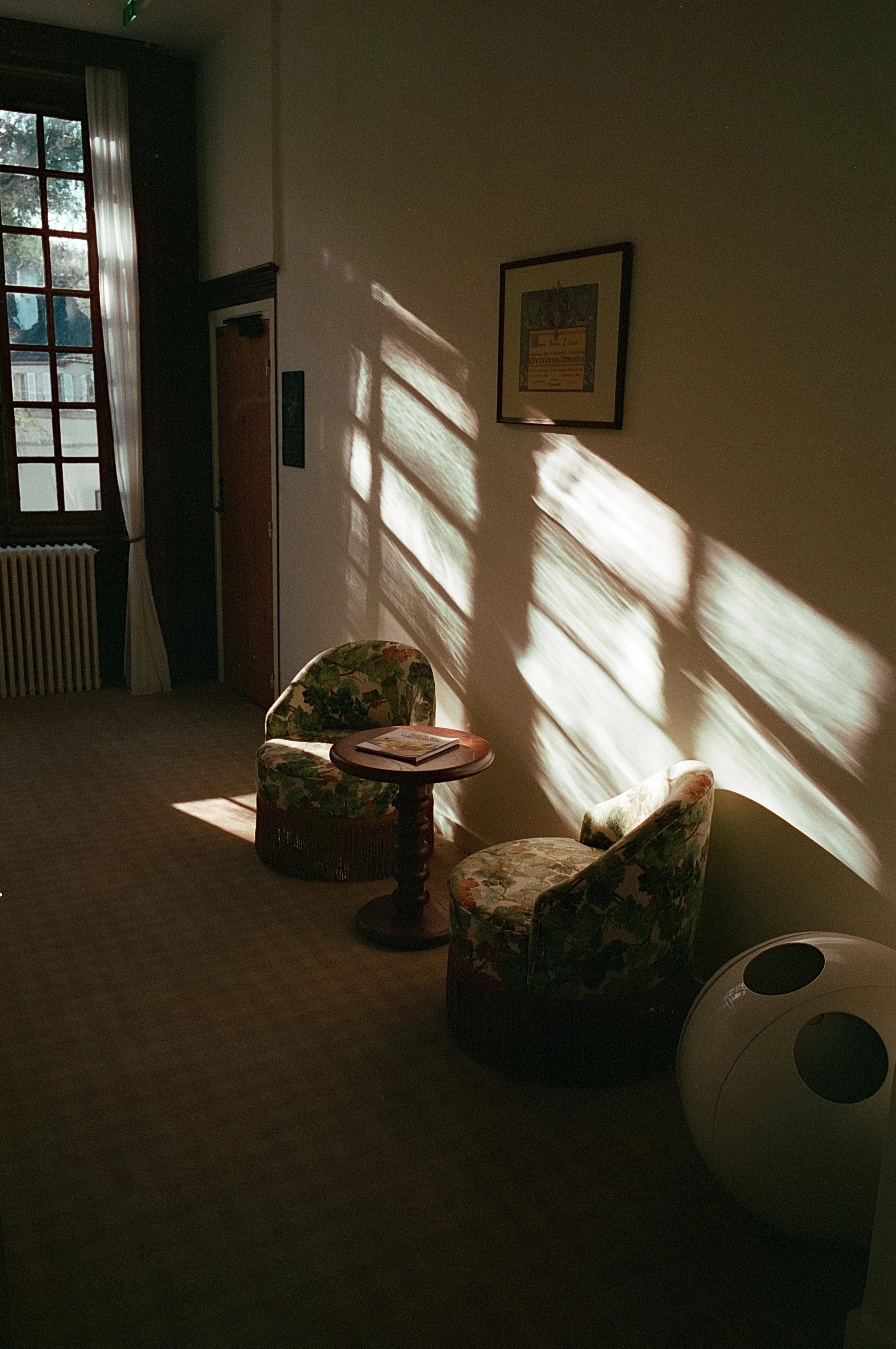 Une pièce éclairée par la lumière du soleil qui filtre à travers une fenêtre, projetant des ombres sur le mur. Il y a deux fauteuils avec un motif floral, une petite table en bois entre eux avec un magazine dessus, et un appareil de purification d'ai