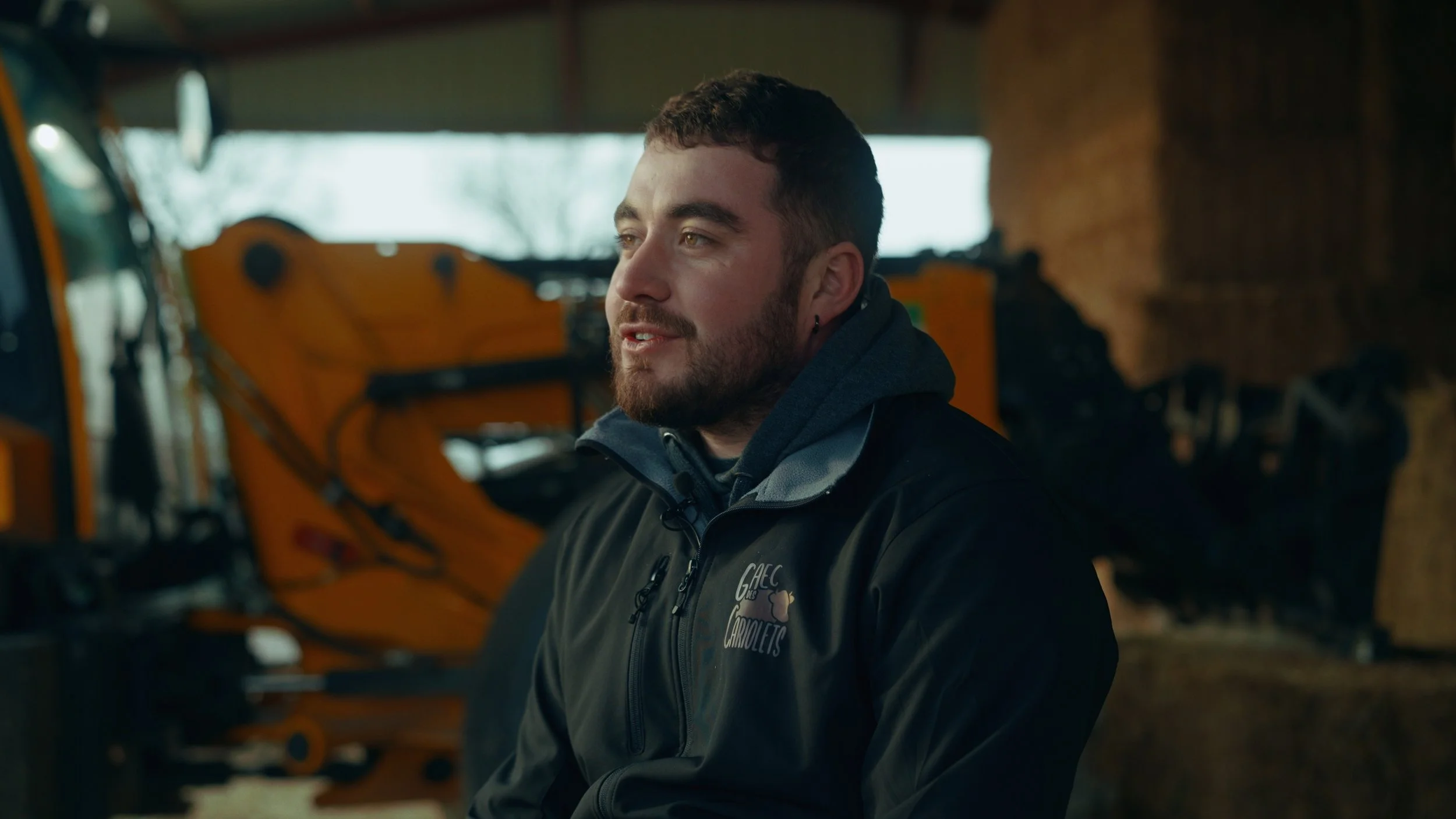 Jeune homme avec barbe, portant un manteau noir avec une capuche, assis dans un bâtiment en bois avec une machine orange en arrière-plan, regardant vers la droite.