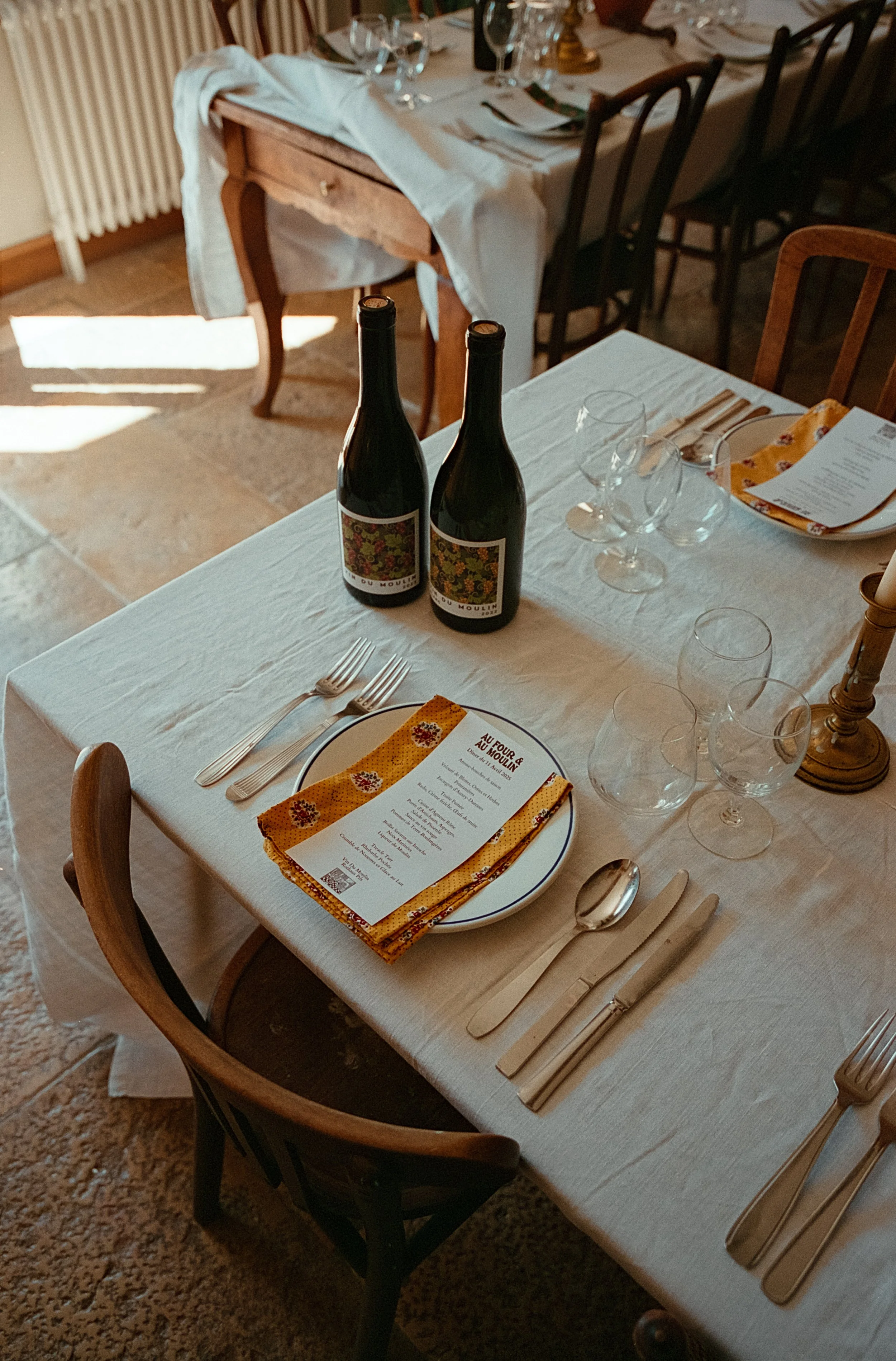 Table dressée avec deux bouteilles de vin, plusieurs verres à vin, des couverts, une assiette avec une nappe en tissu jaune à motif floral, un menu, une bougie et une nappe blanche.