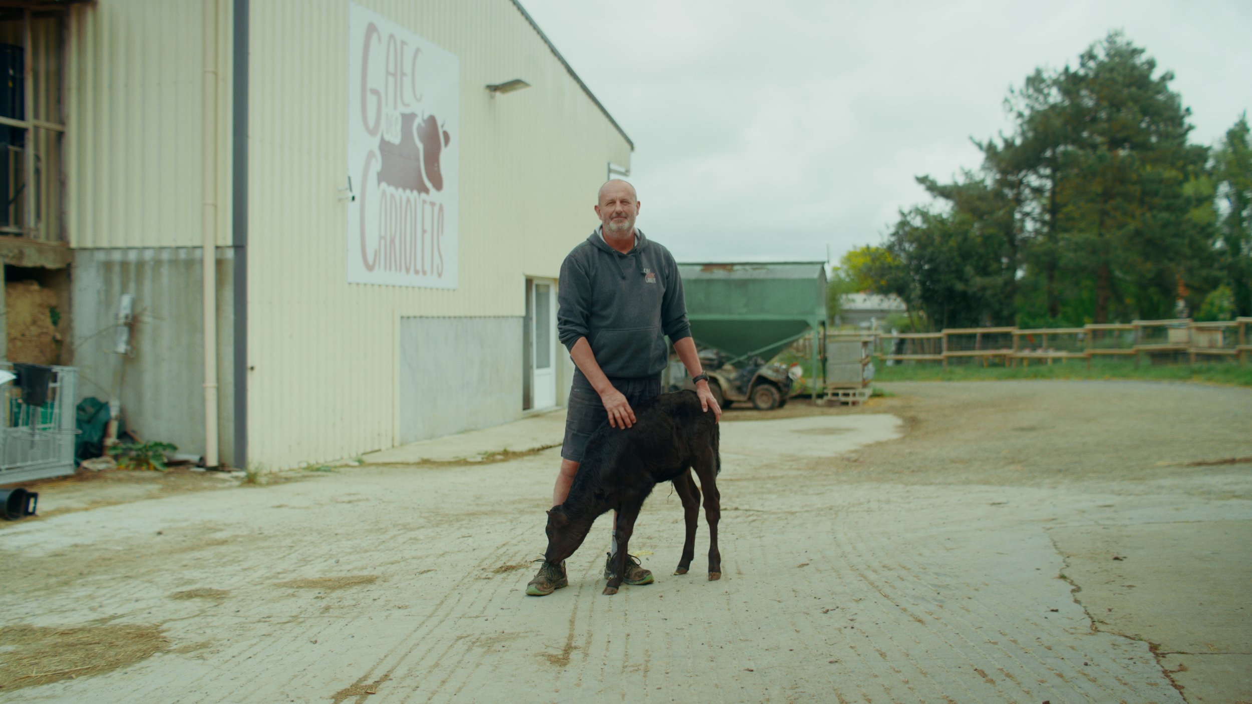 Un homme debout à côté d'un veau noir sur une ferme, en arrière-plan un bâtiment avec une enseigne 'GAEC Charlotte'.