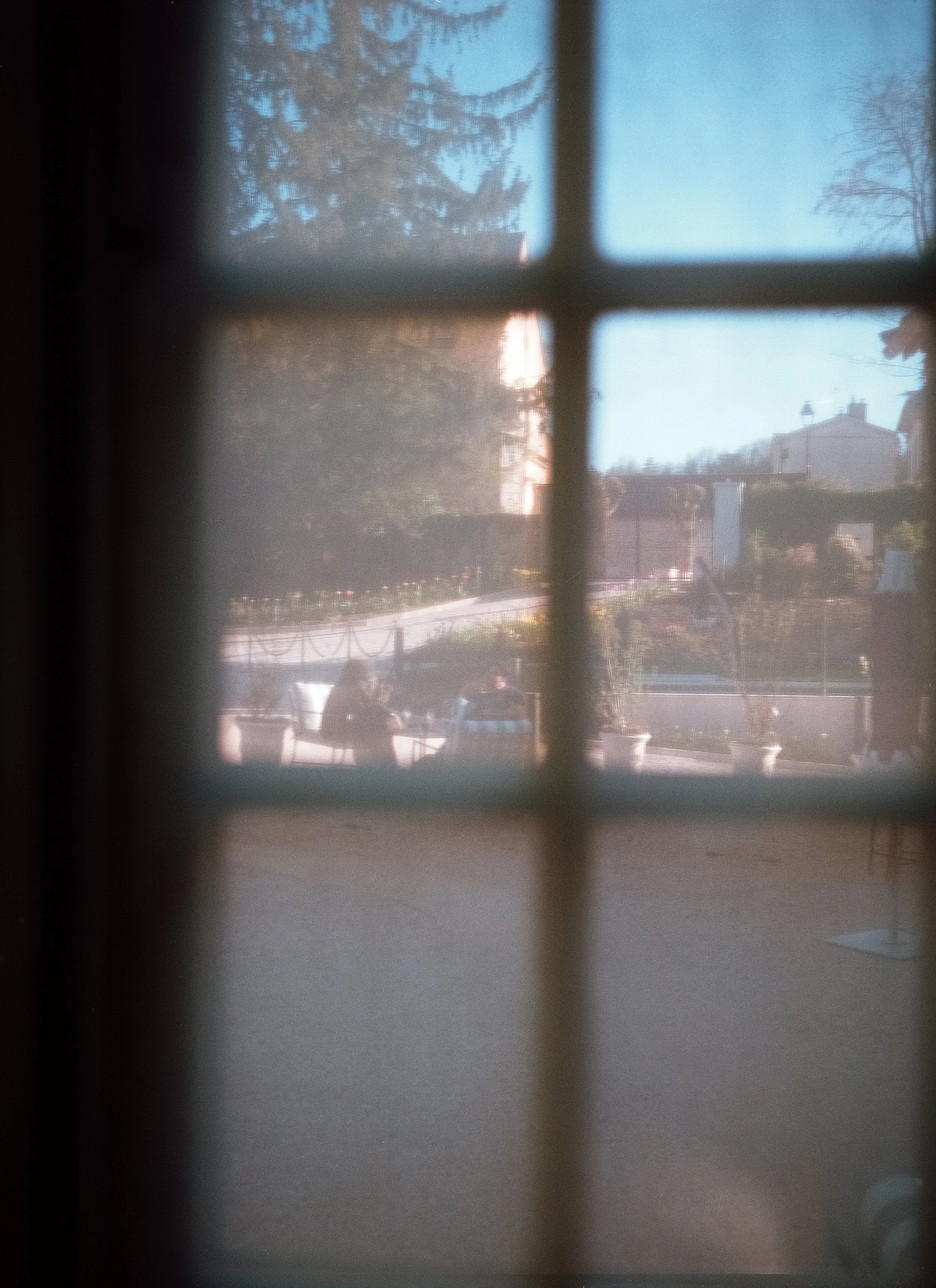 Vue floue à travers une fenêtre en verre dépoli montrant des personnes assises à une table en plein air, avec des bâtiments et des arbres au loin dans un ciel bleu.