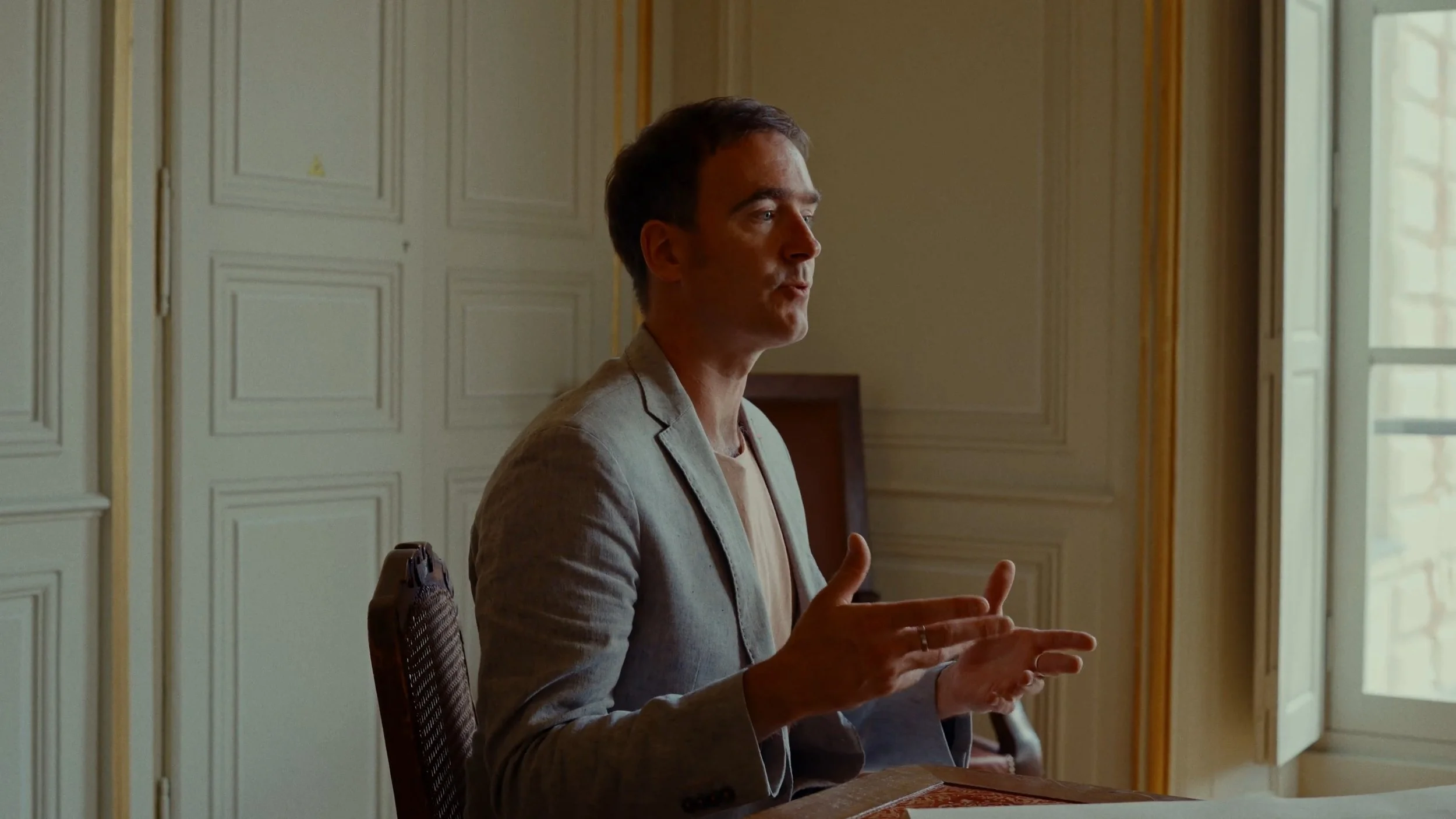 Un homme en costume beige parle lors d'une réunion dans une pièce avec des murs ornés de moulures, près d'une fenêtre laissant passer la lumière