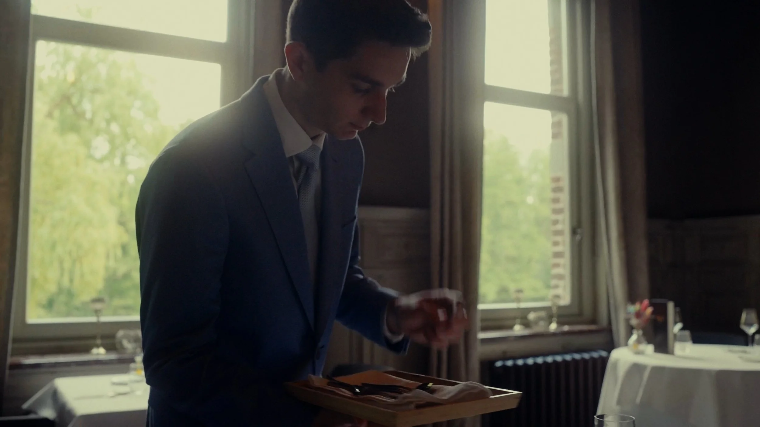 Jeune homme en costume dans une salle à manger, tenant un plateau avec des couverts, aux fenêtres avec rideaux, ambiance naturelle et élégante.
