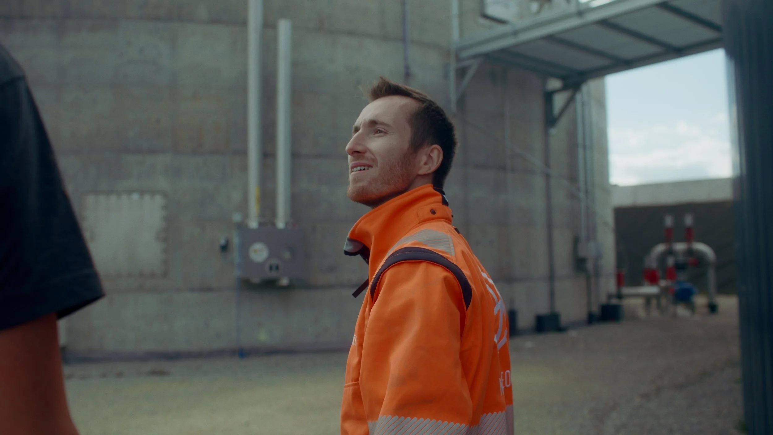 Jeune homme portant une veste orange dans une usine ou un entrepôt, souriant et regardant vers le haut.