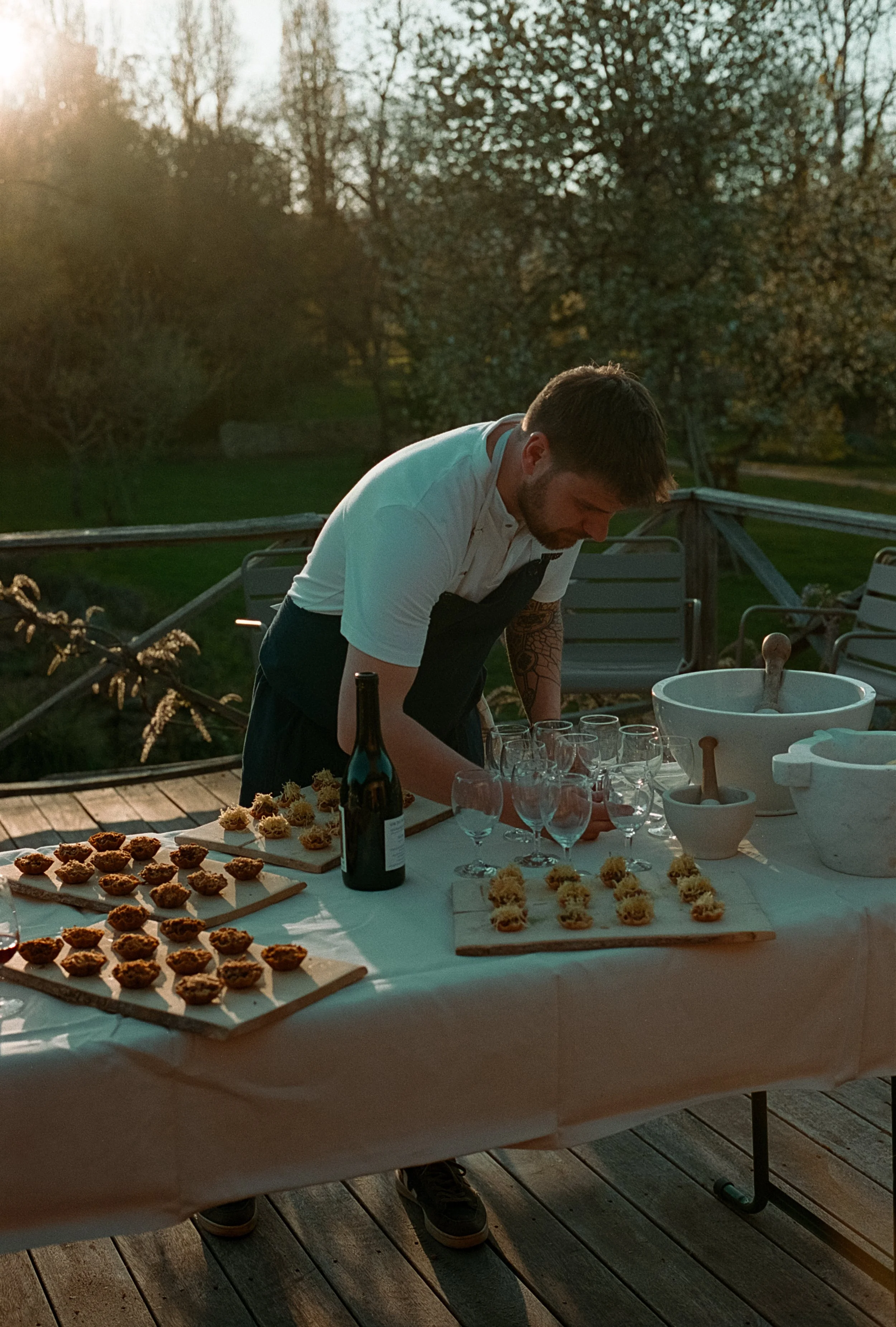 Un homme prépare des verres à proximité d'amuse-bouches sur une table lors d'un coucher de soleil en extérieur.
