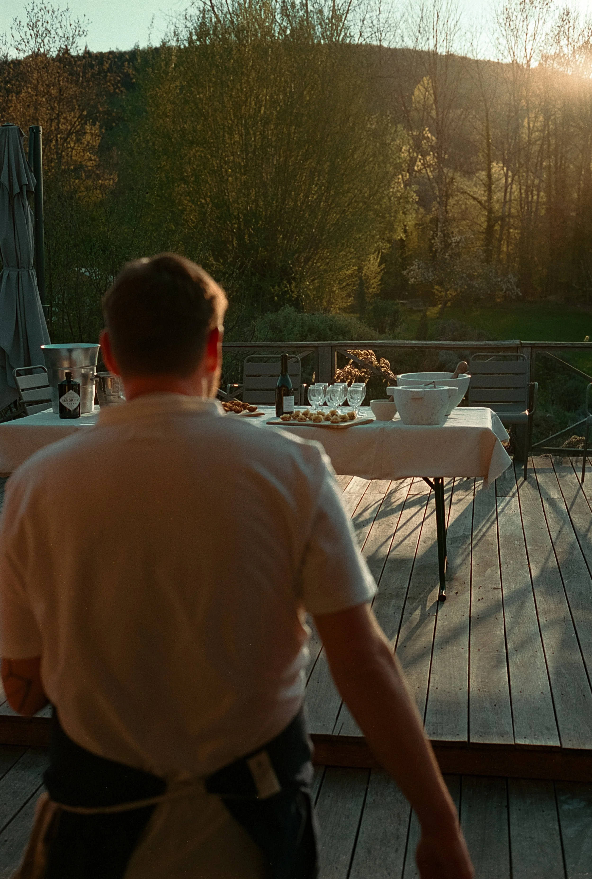 Un homme vu de dos, portant un tablier blanc, se tient sur une terrasse en bois. En face de lui, une table est dressée avec des verres, des assiettes et des plats, face à un paysage naturel avec des arbres et un coucher de soleil.