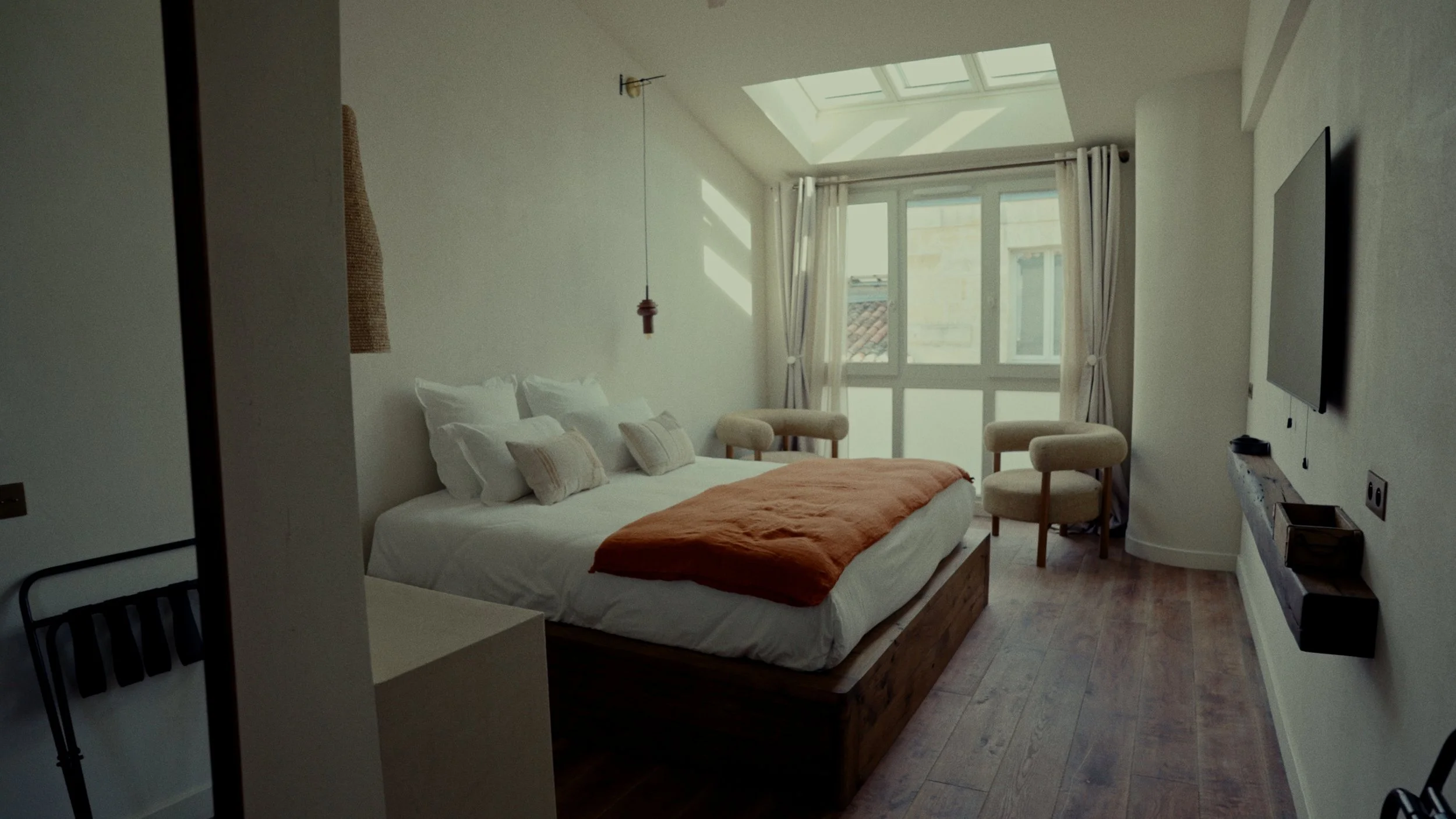 Chambre avec grand lit à baldaquin, conçue dans des tons neutres, avec fenêtres lumineuses, fauteuils, et un téléviseur mural.