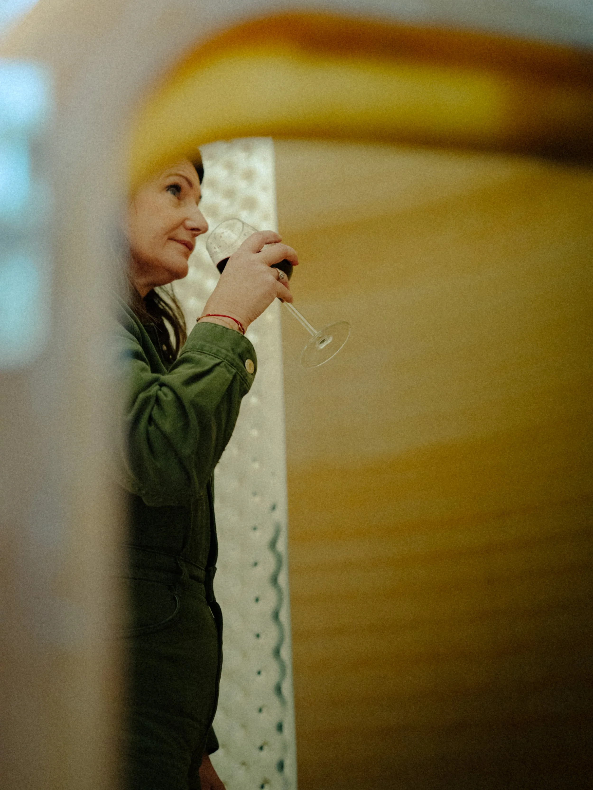 Une femme en pull vert sombre tenant un verre de vin rouge, vue de côté, en train de boire.