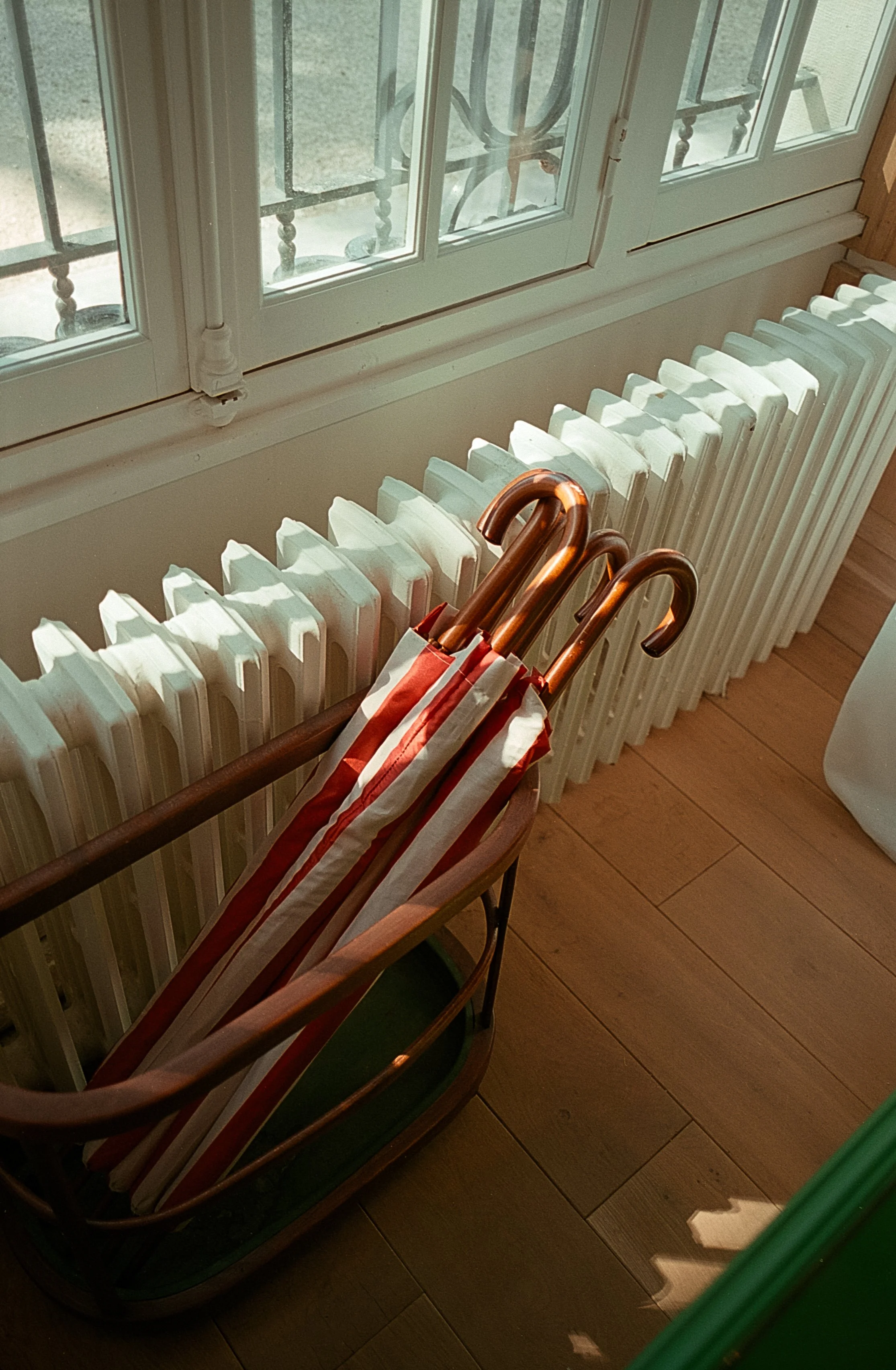 Un porte-parapluies en bois avec plusieurs parapluies rouges et blancs à l'intérieur, placé près d'une radiateur en métal blanc au sol, avec une fenêtre en arrière-plan.