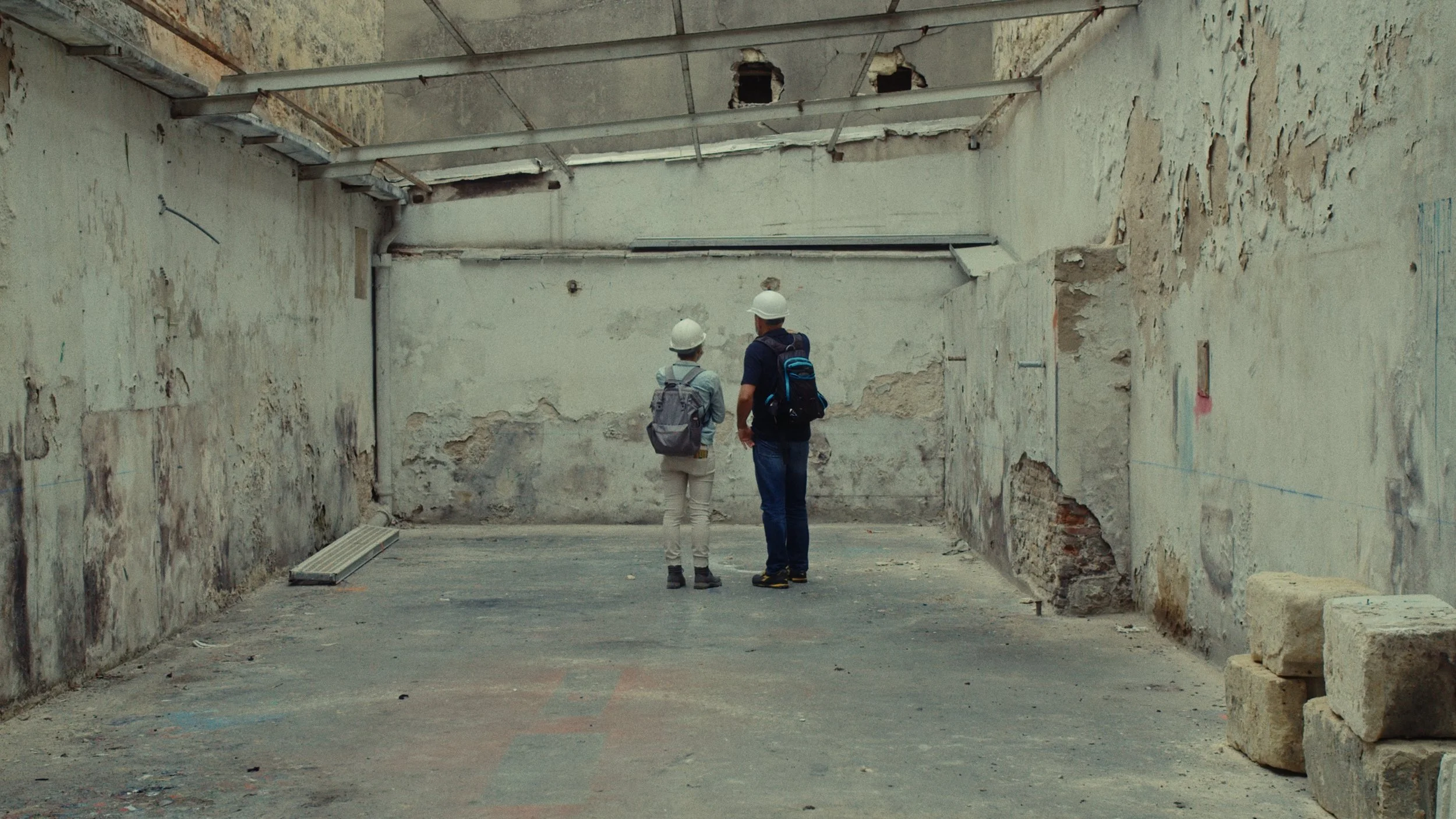 Deux personnes portant des casques de sécurité et des sacs à dos, se tenant dans une pièce en rénovation avec des murs endommagés et dégradés.