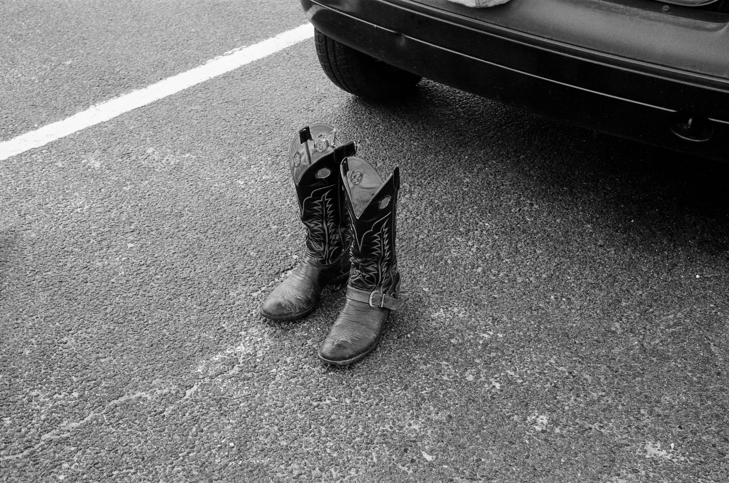 Une paire de bottes de cow-boy est posée sur le sol d'un parking à côté d'une voiture. L'image est en noir et blanc.