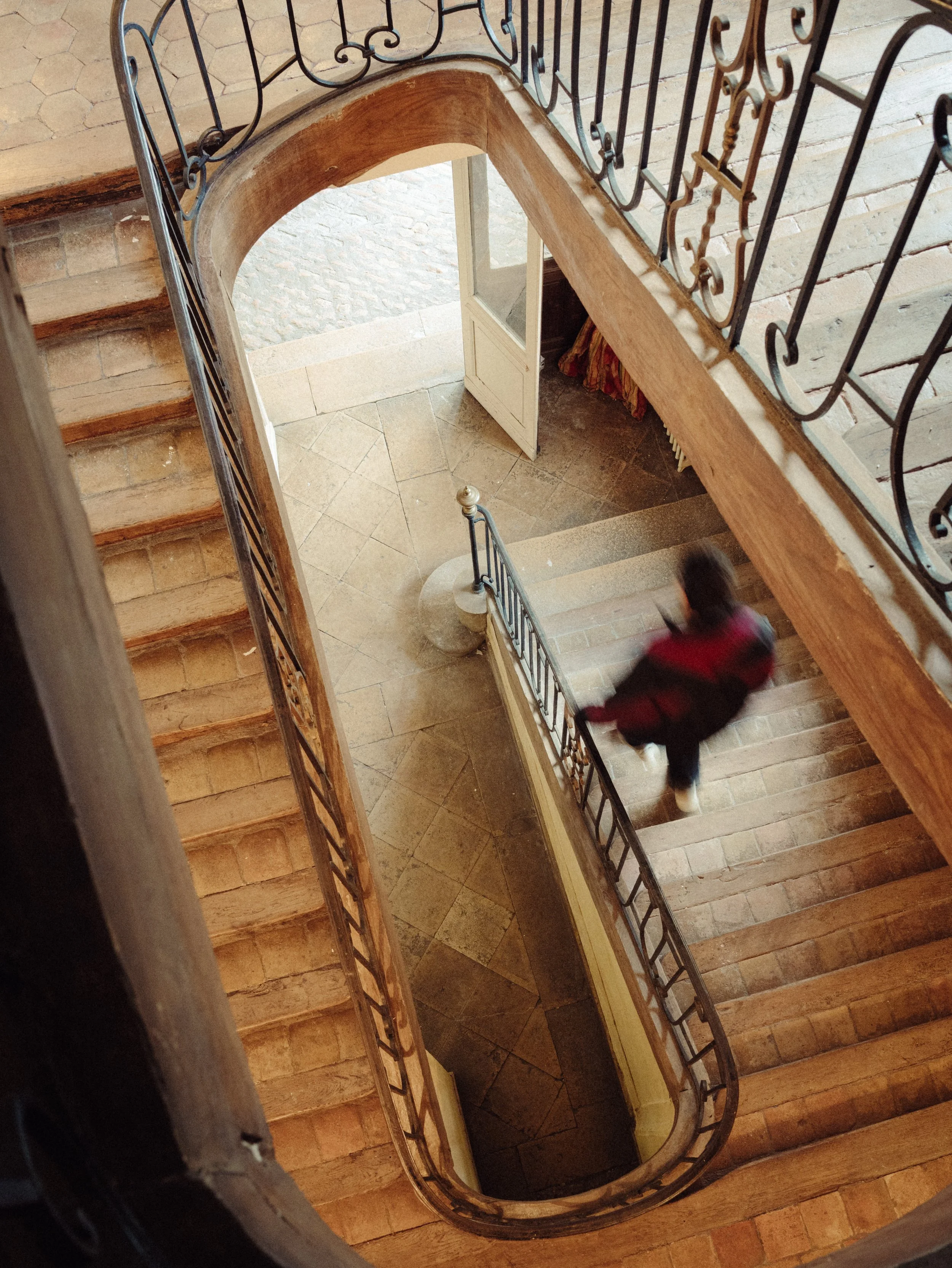 Vue d'un escalier en bois avec des rampes en métal, vu du dessus. Un personnage flou, une femme, descend l'escalier. À l'étage, on voit une porte vitrée ouverte et un sol en pierre.