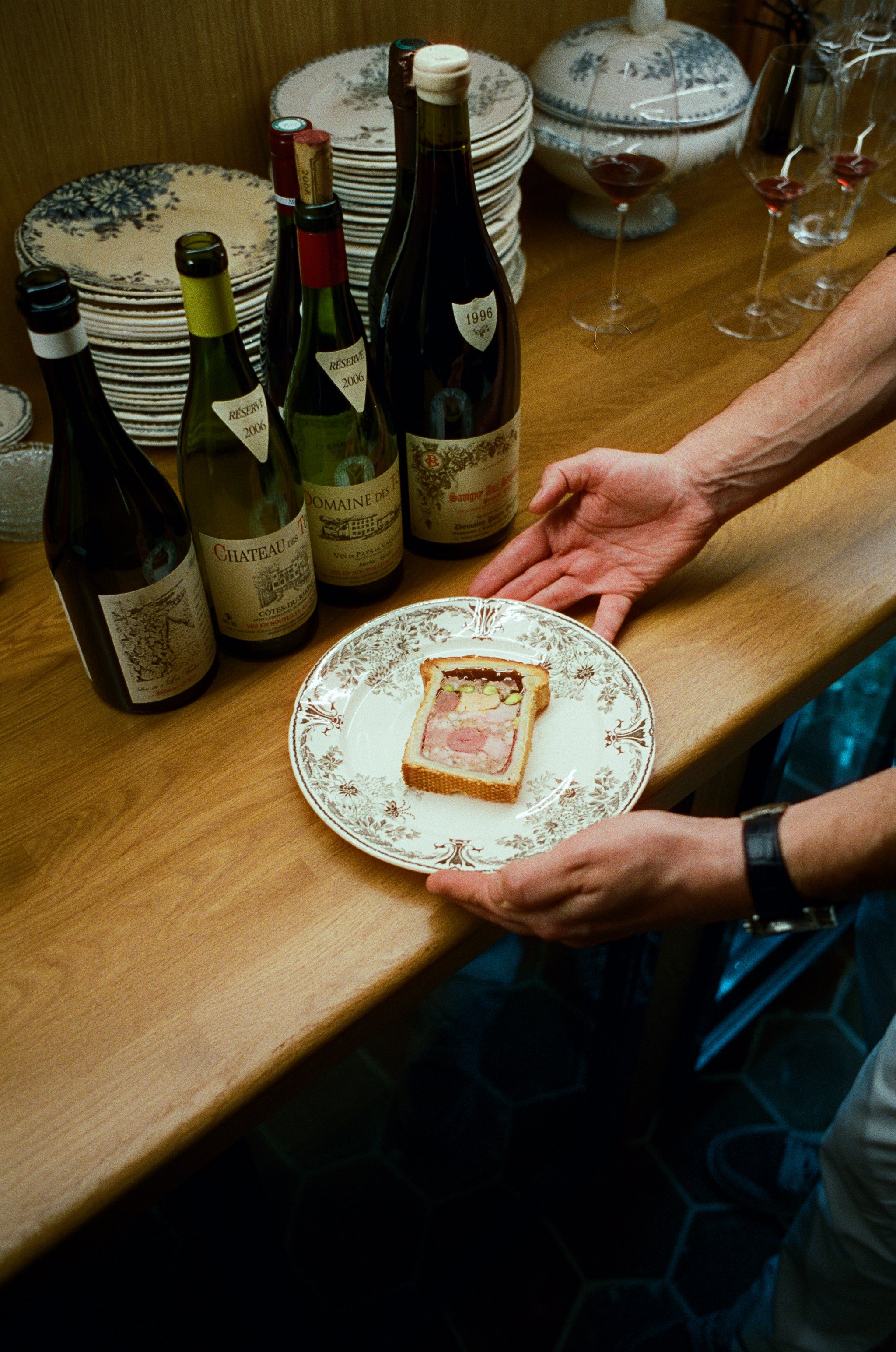 Personne tenant une assiette avec une tranche de pâté en croûte devant une rangée de bouteilles de vin.