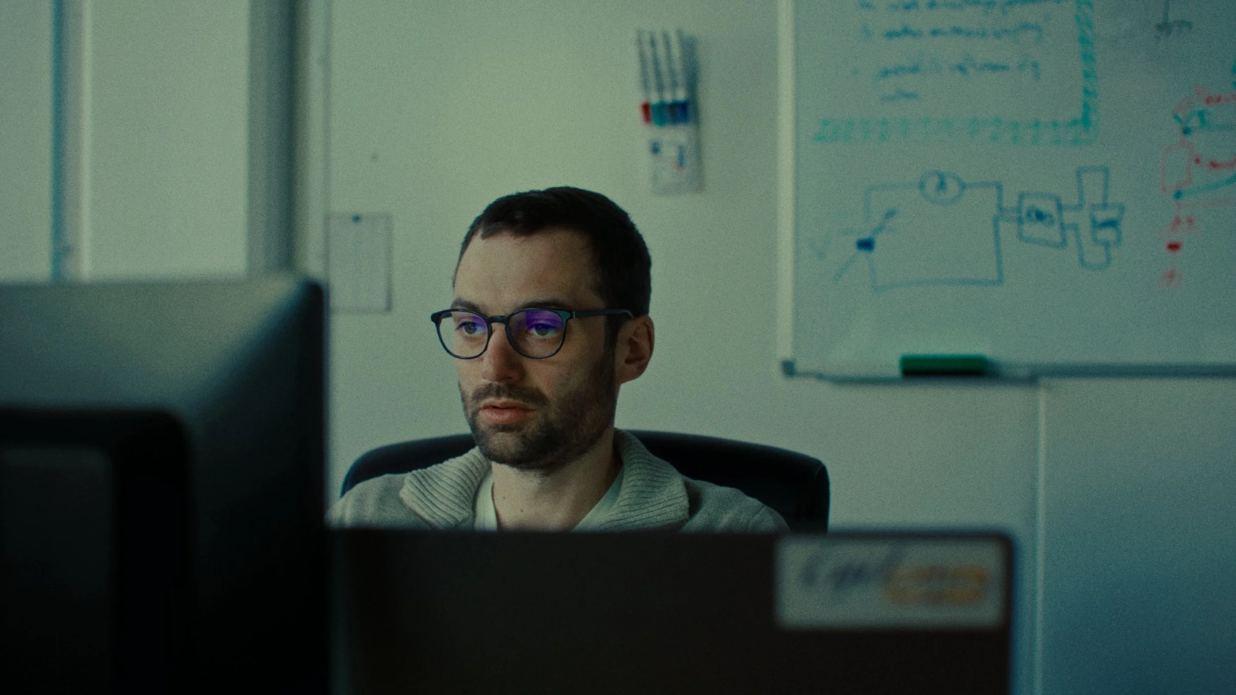 Un homme portant des lunettes assis à un bureau devant un ordinateur, avec un tableau blanc derrière lui contenant des schémas et des notes.