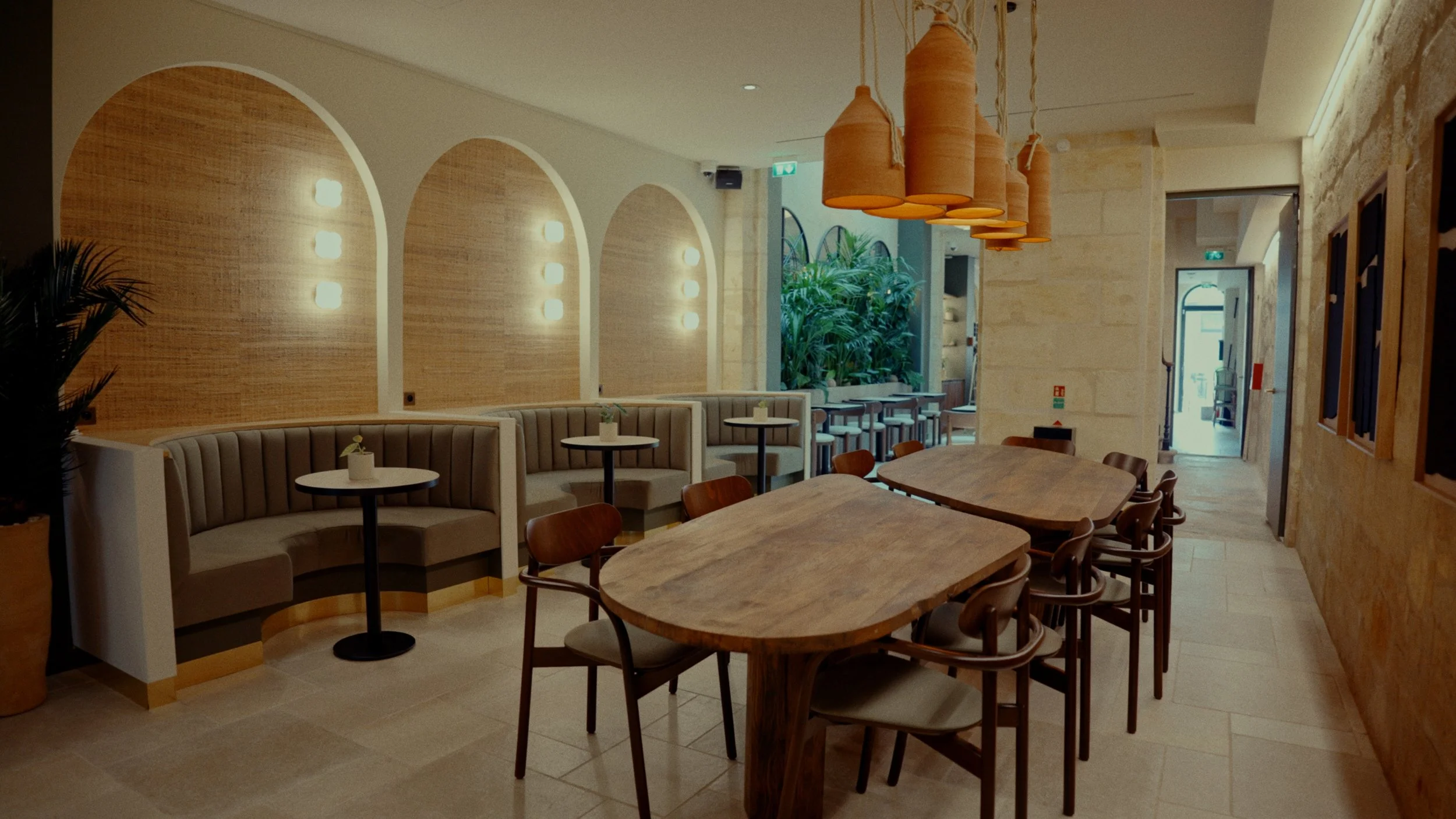 Intérieur d'un café ou restaurant avec mobilier en bois, banquettes beige, tables rondes et rectangulaires, suspension en terre cuite, murs en pierre et végétation visible à l'arrière.