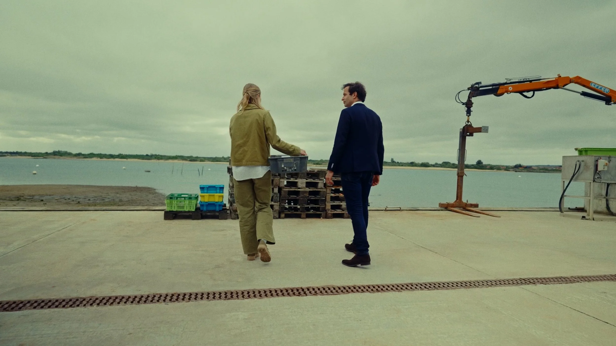 Deux personnes, un homme en costume et une femme en tenue décontractée, discutent près d'une étendue d'eau, avec des palettes en bois et des caisses en plastique autour, sous un ciel nuageux.