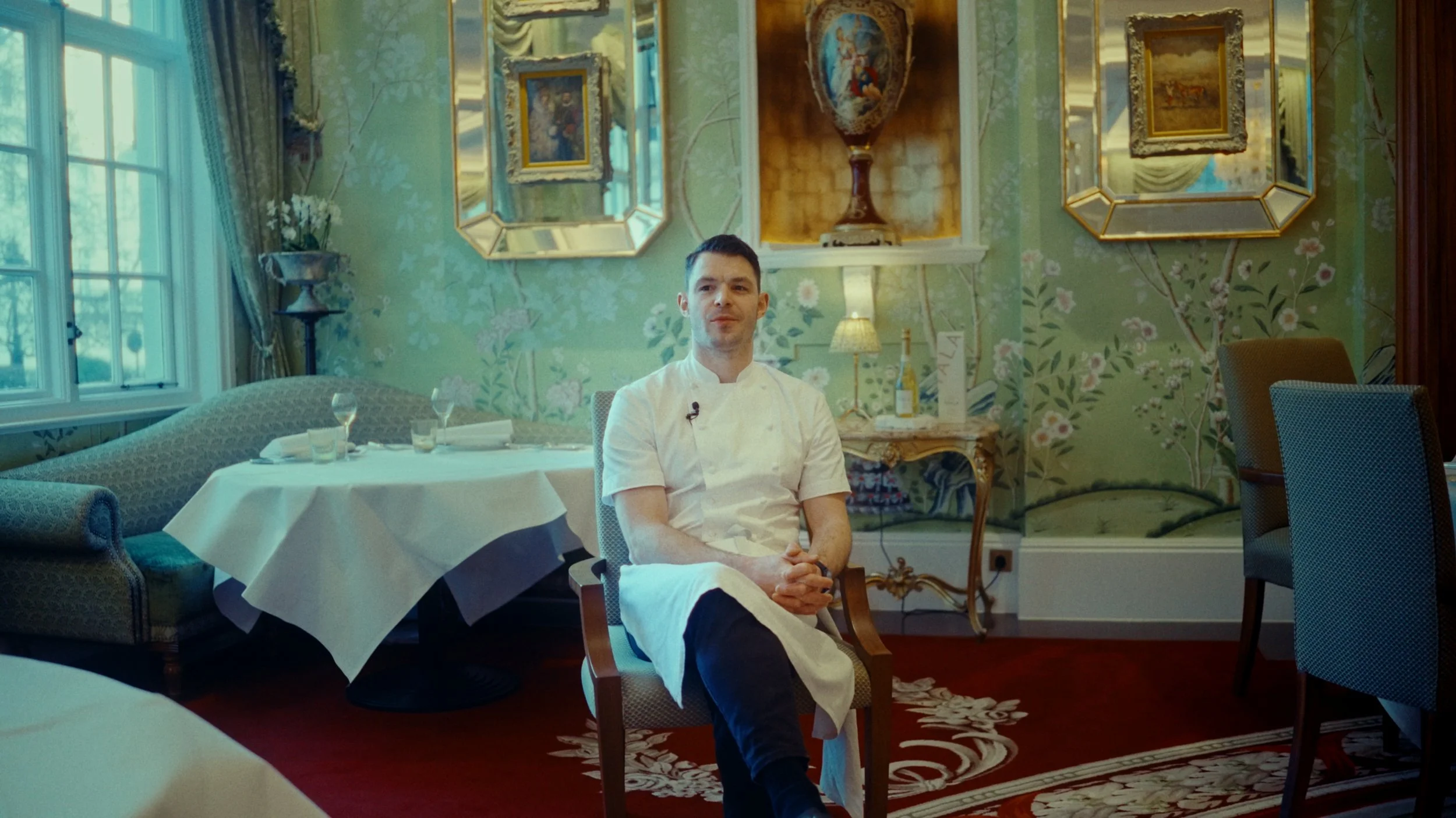 Un jeune chef en uniforme blanc assis dans un salon élégant, avec des tableaux et des miroirs dorés, une fenêtre avec des rideaux à motif floral, une table entretenue et des décorations classiques