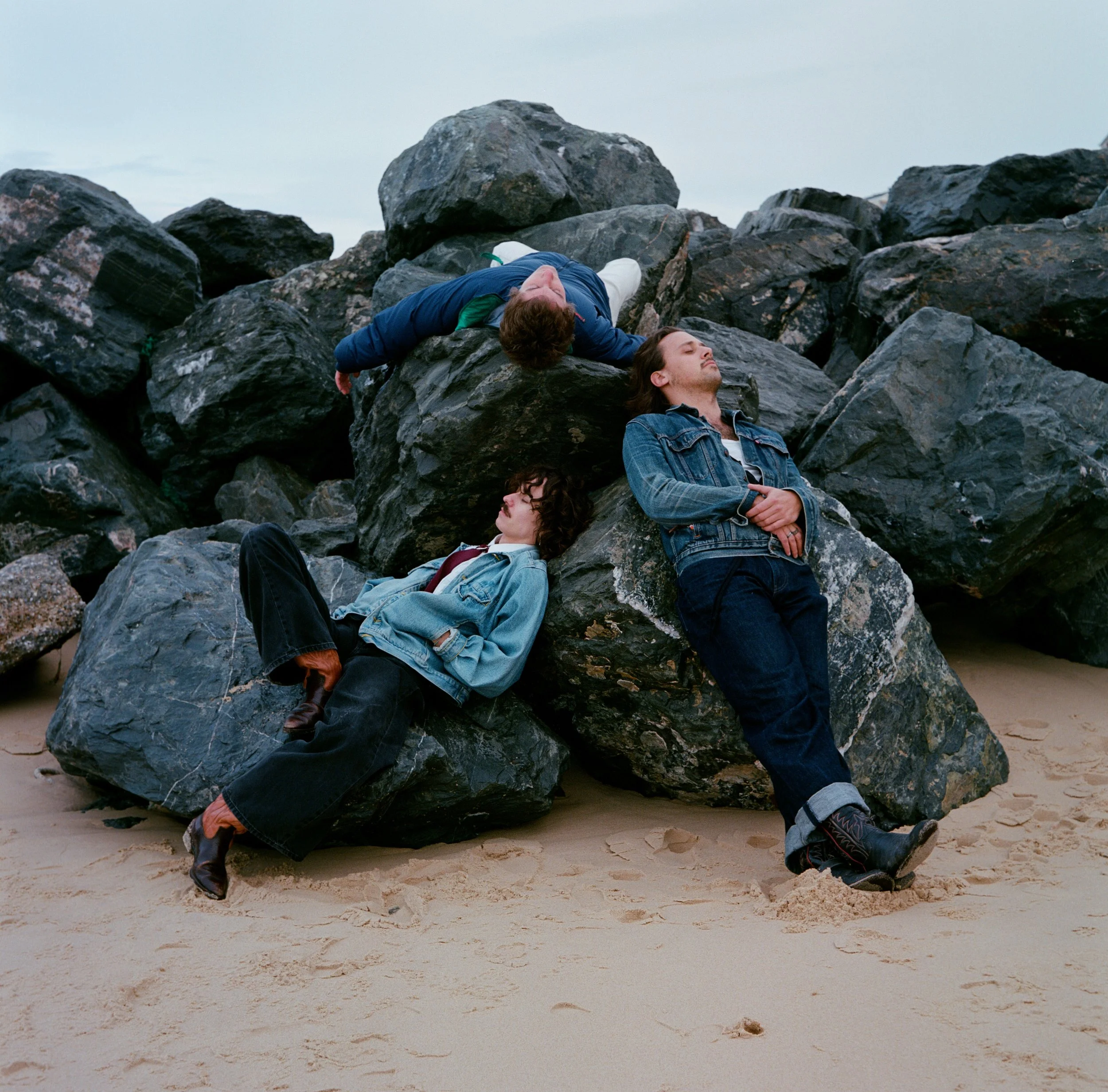 Trois jeunes hommes en vêtements décontractés posent sur des gros rochers au bord de la plage, avec un ciel nuageux en arrière-plan.