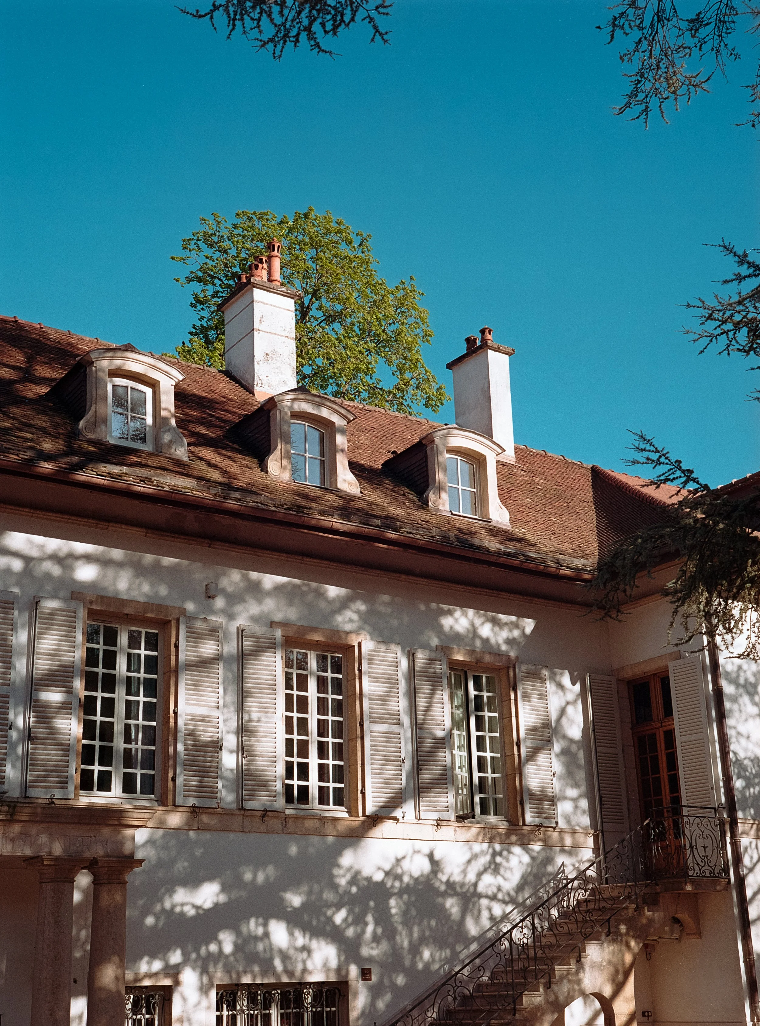 Maison avec volets blancs, fenêtres à petits carreaux, toit en tuiles, deux cheminées, et escaliers extérieurs, sous un ciel bleu, avec des arbres en ombre chinoise.