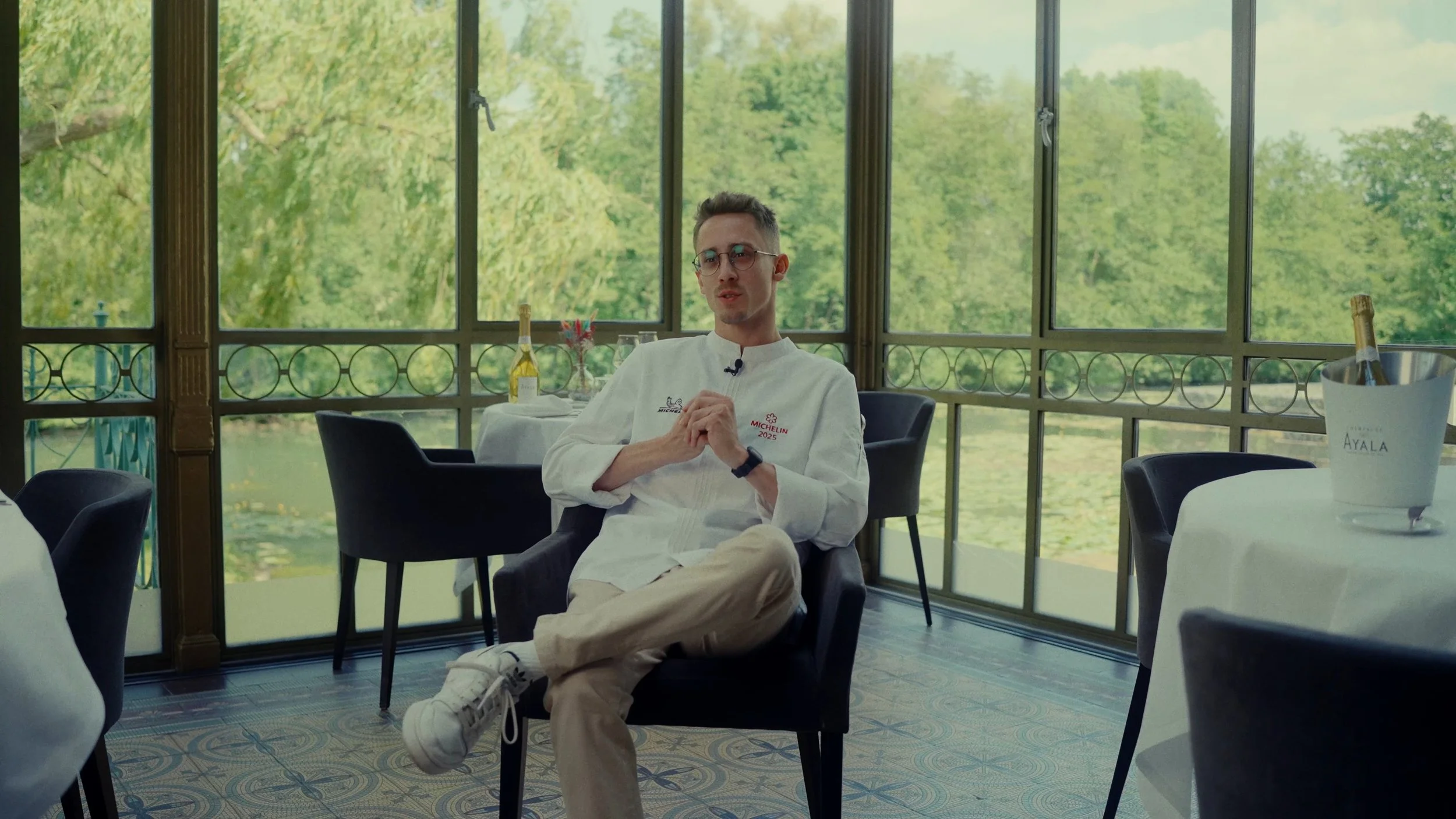 Un homme habillé en chef cuisinier, portant un costume blanc avec des logos Michelin et Michelin 2023, assis dans une salle avec vue sur un lac et des arbres, avec une bouteille de champagne dans un seau à glace sur une table à côté.