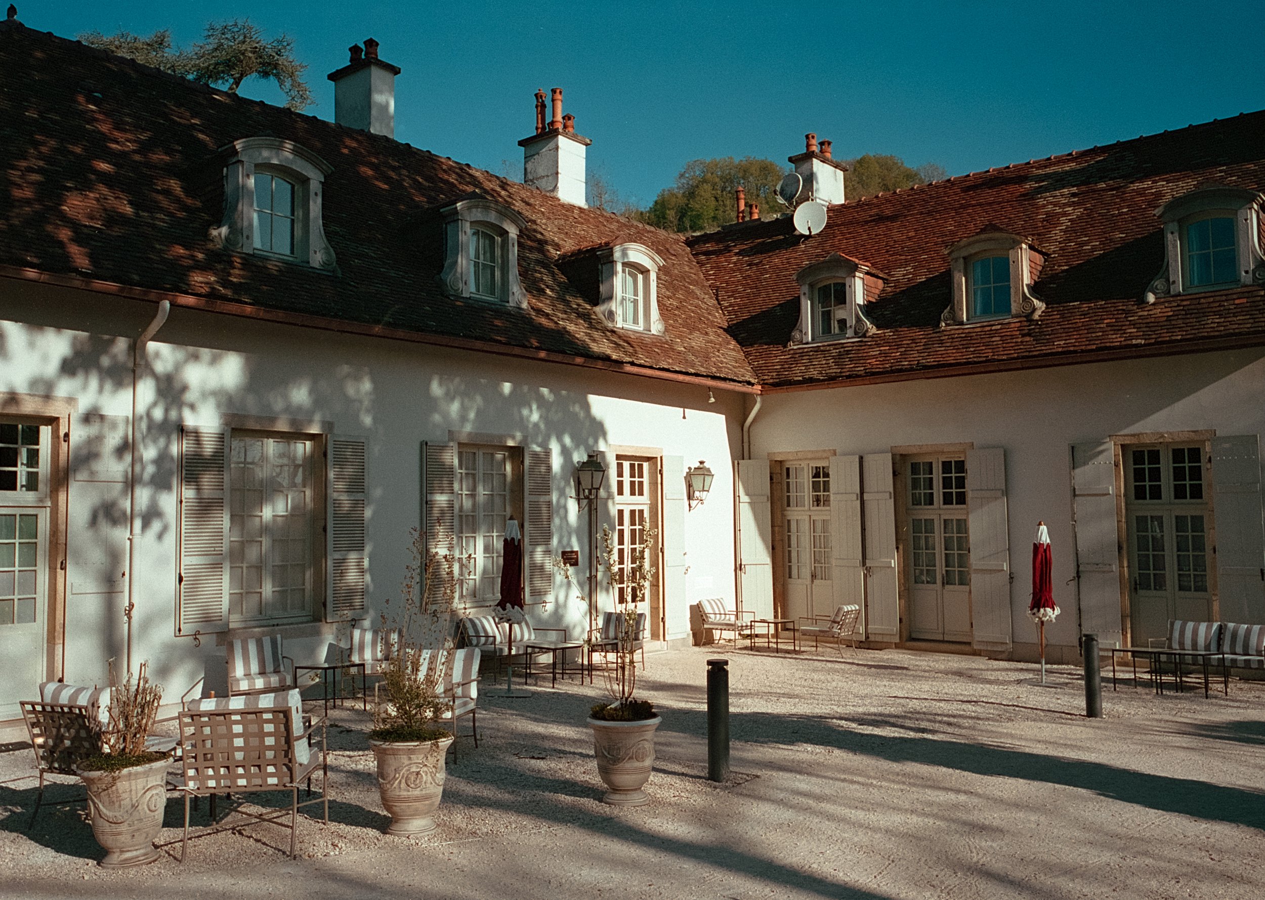 Cour intérieure d'une maison avec plusieurs fenêtres à volets, chaises, tables, et parasols rouges, sous un ciel bleu.
