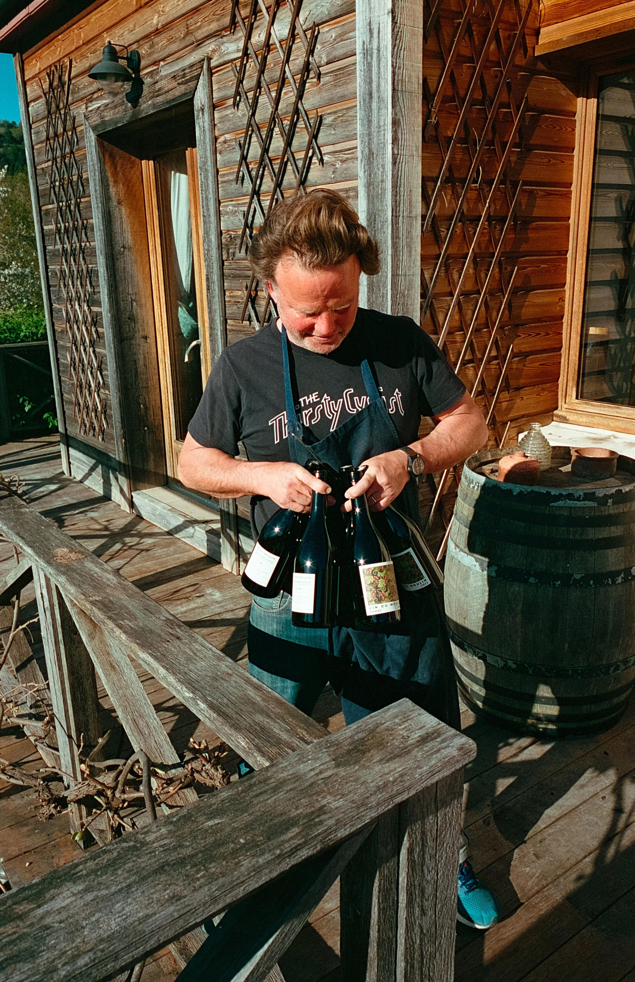 Un homme tient une poignées de bouteilles de vin, debout sur une terrasse en bois devant une maison en bois.