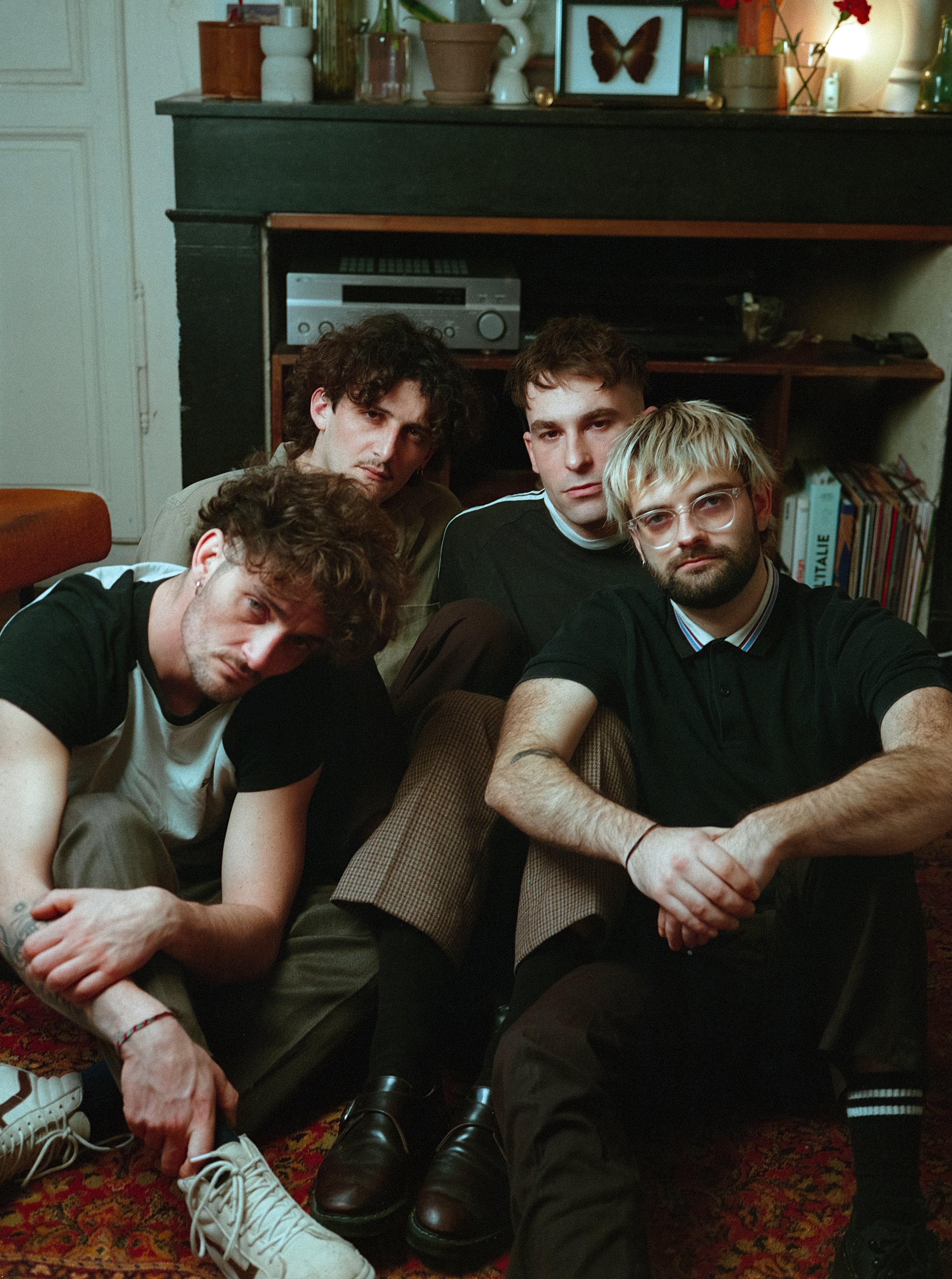 Un groupe de cinq jeunes hommes posant dans un salon, assis ou accroupis, avec une ambiance décontractée et un style vintage.