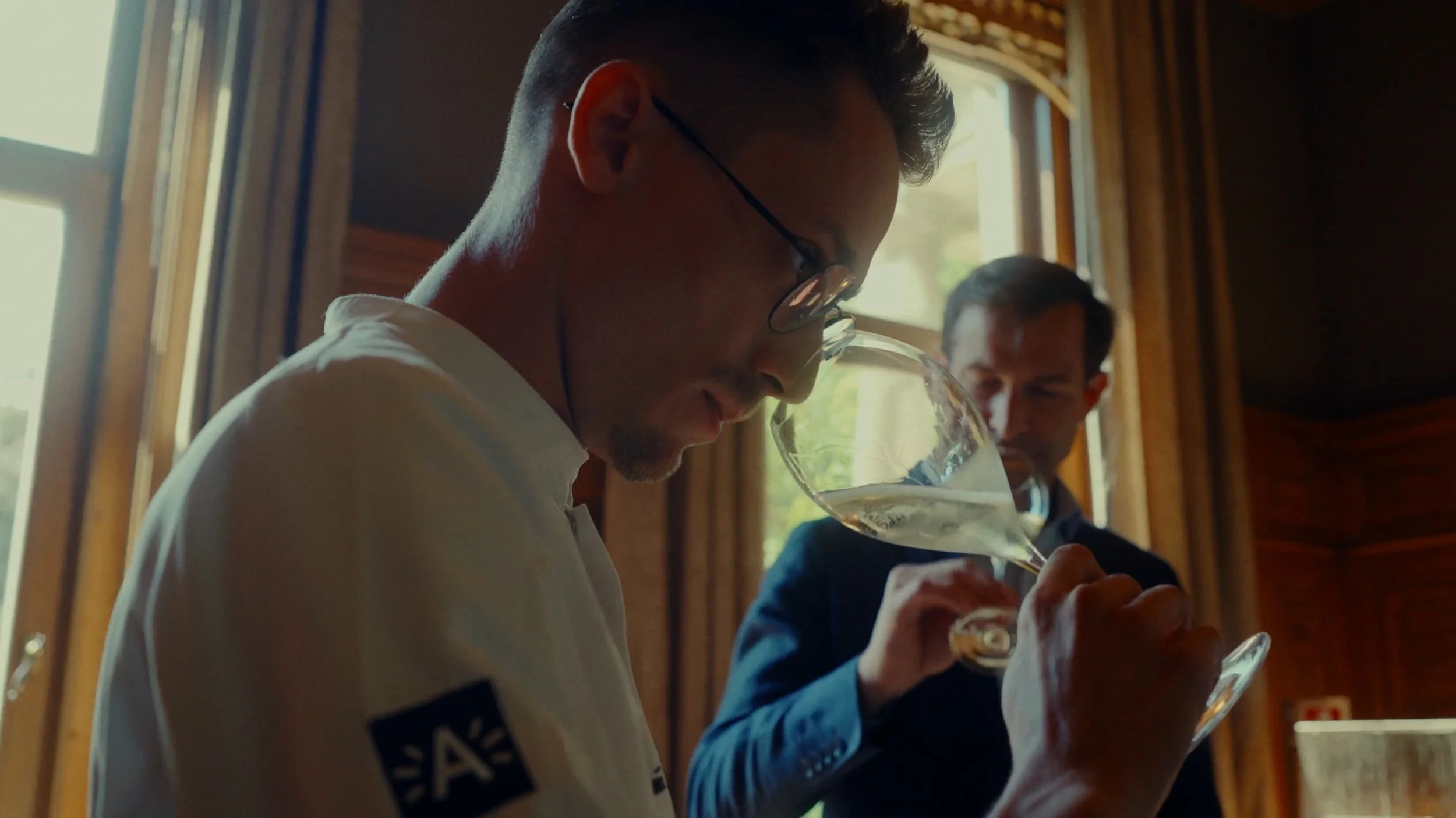 Deux hommes dégustent du vin dans une pièce avec des murs en bois et des fenêtres laissant entrer la lumière naturelle.