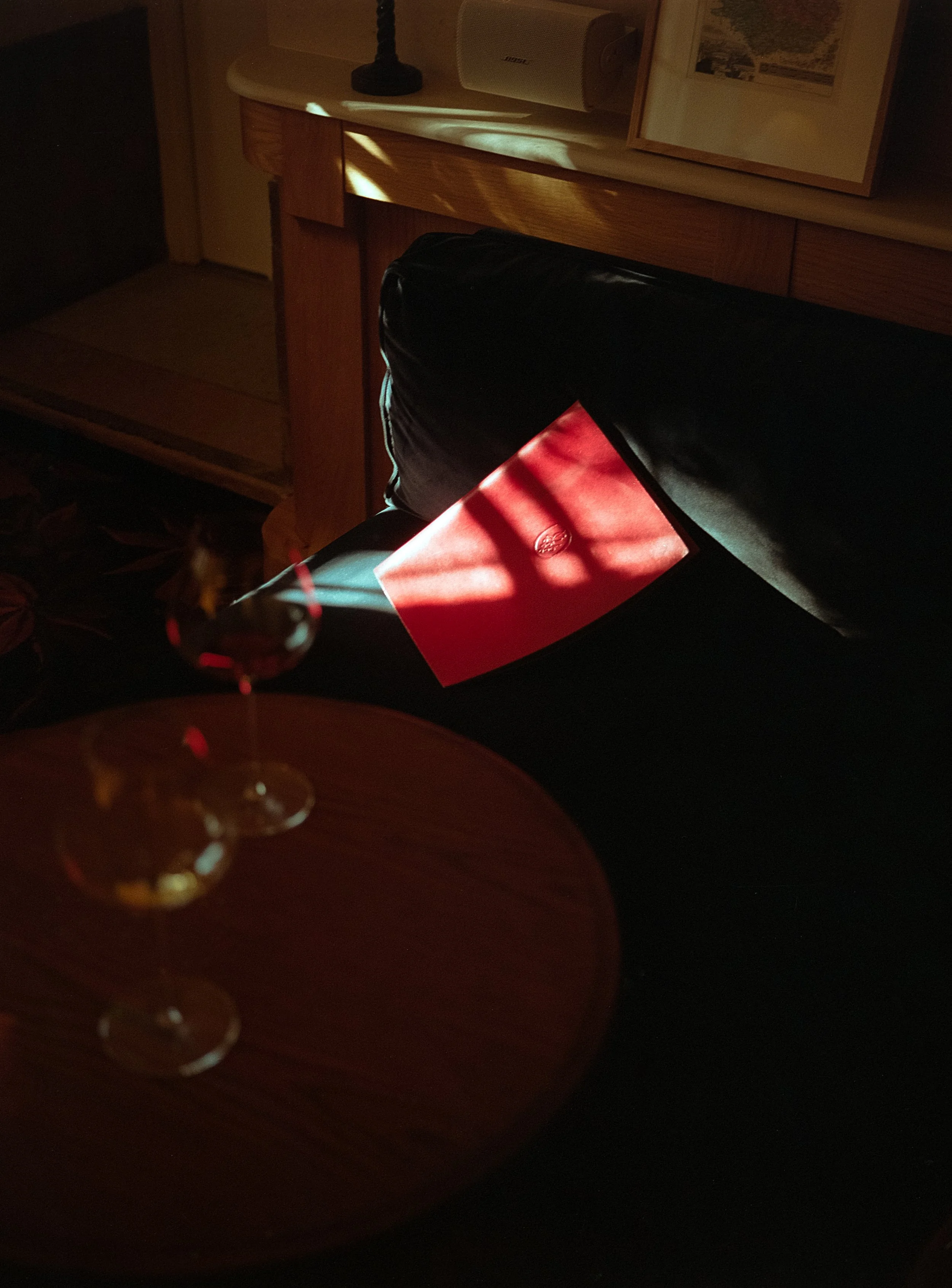 Une pièce sombre avec une table en bois ronde, deux verres à vin, une chaise noire avec un coussin, un meuble en bois avec une lampe et un cadre photo sur une étagère, une bouteille de vin rouge et un livre rouge posés sur la chaise. La lumière natur