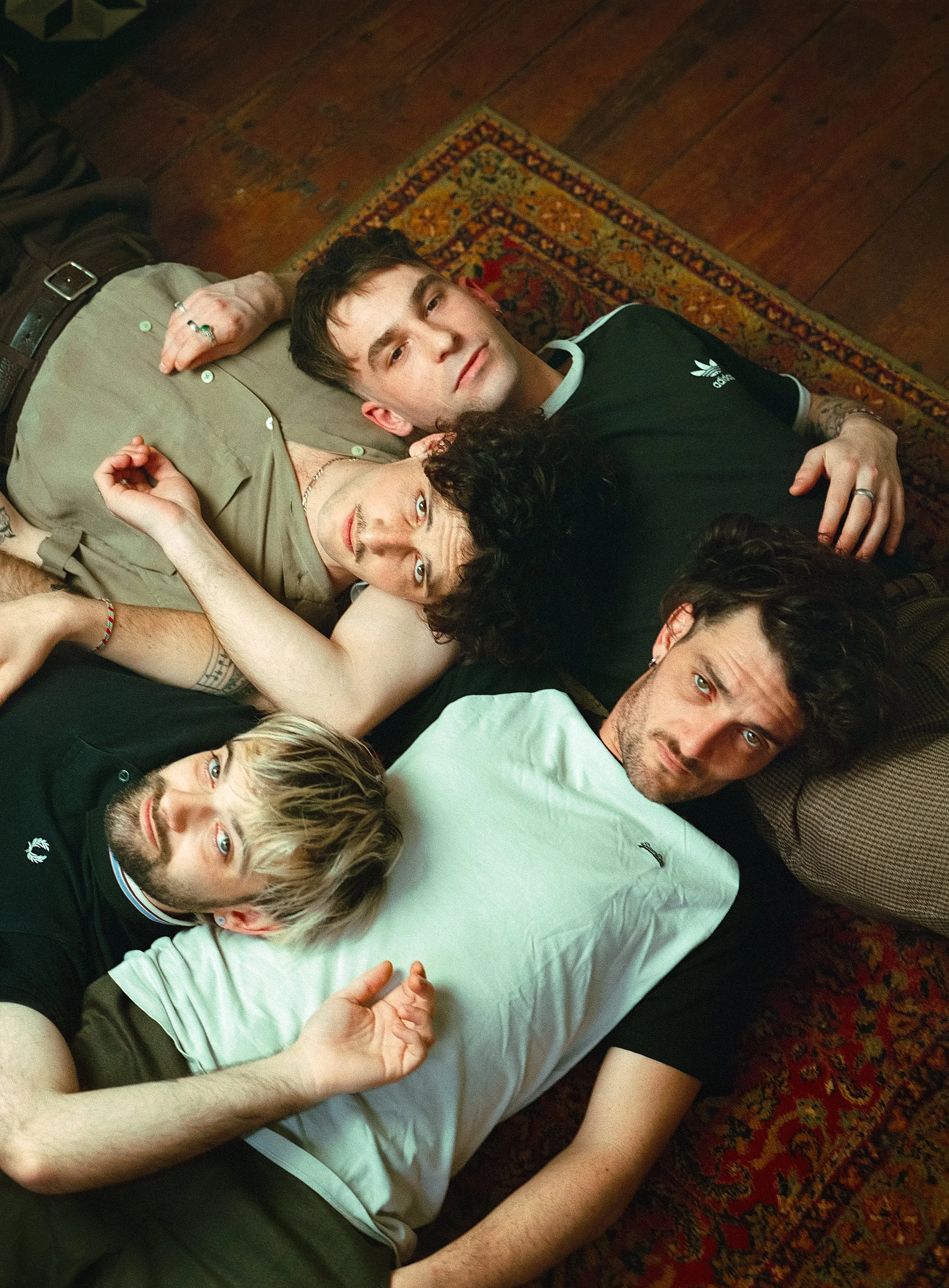 Cinq jeunes hommes allongés sur un tapis, regardant vers l'objectif, certains avec des expressions sérieuses, dans une pièce boisée.