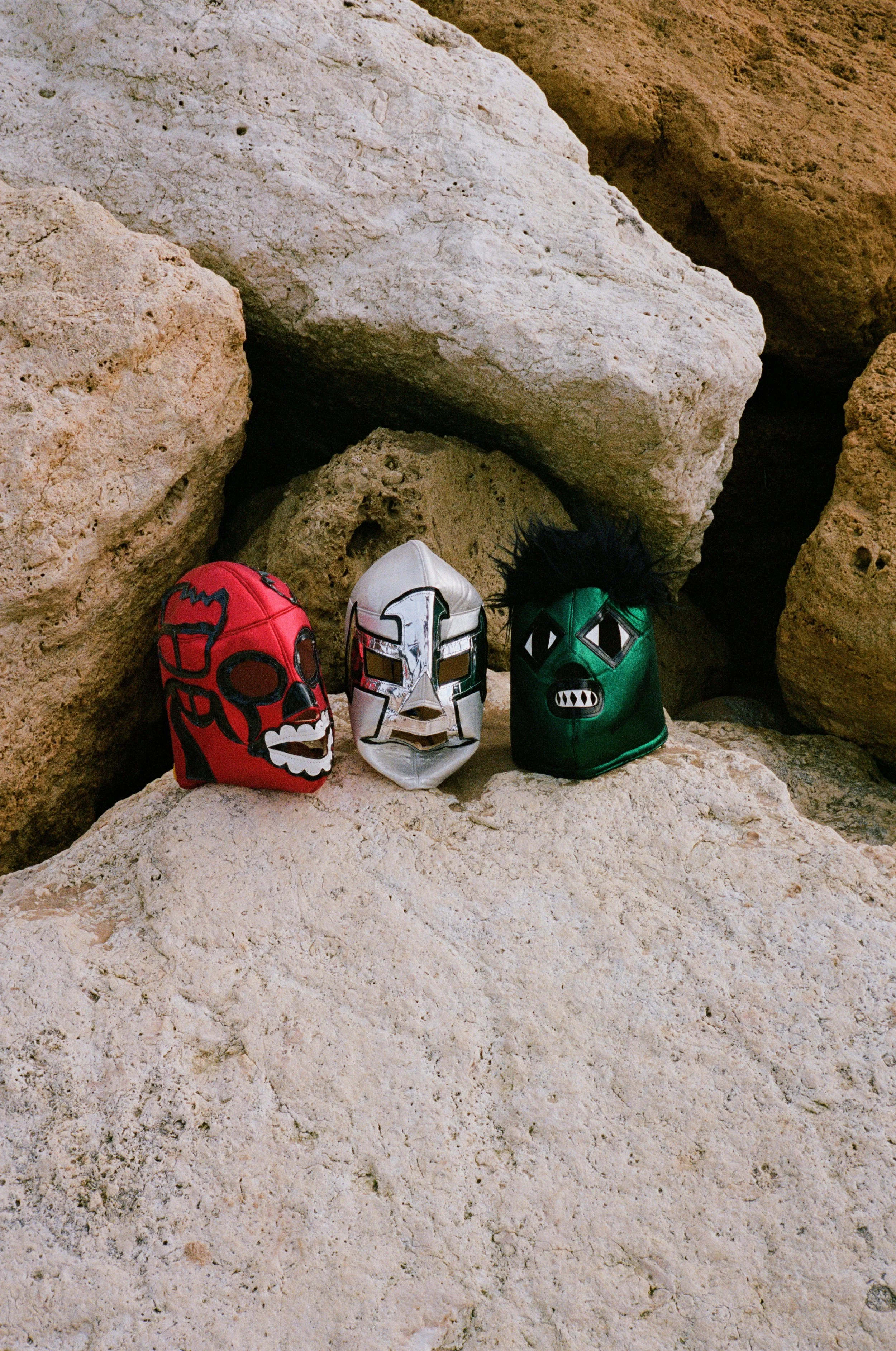 Trois masques de carnaval avec des visages de personnages mythiques, posés sur des rochers rocheux.
