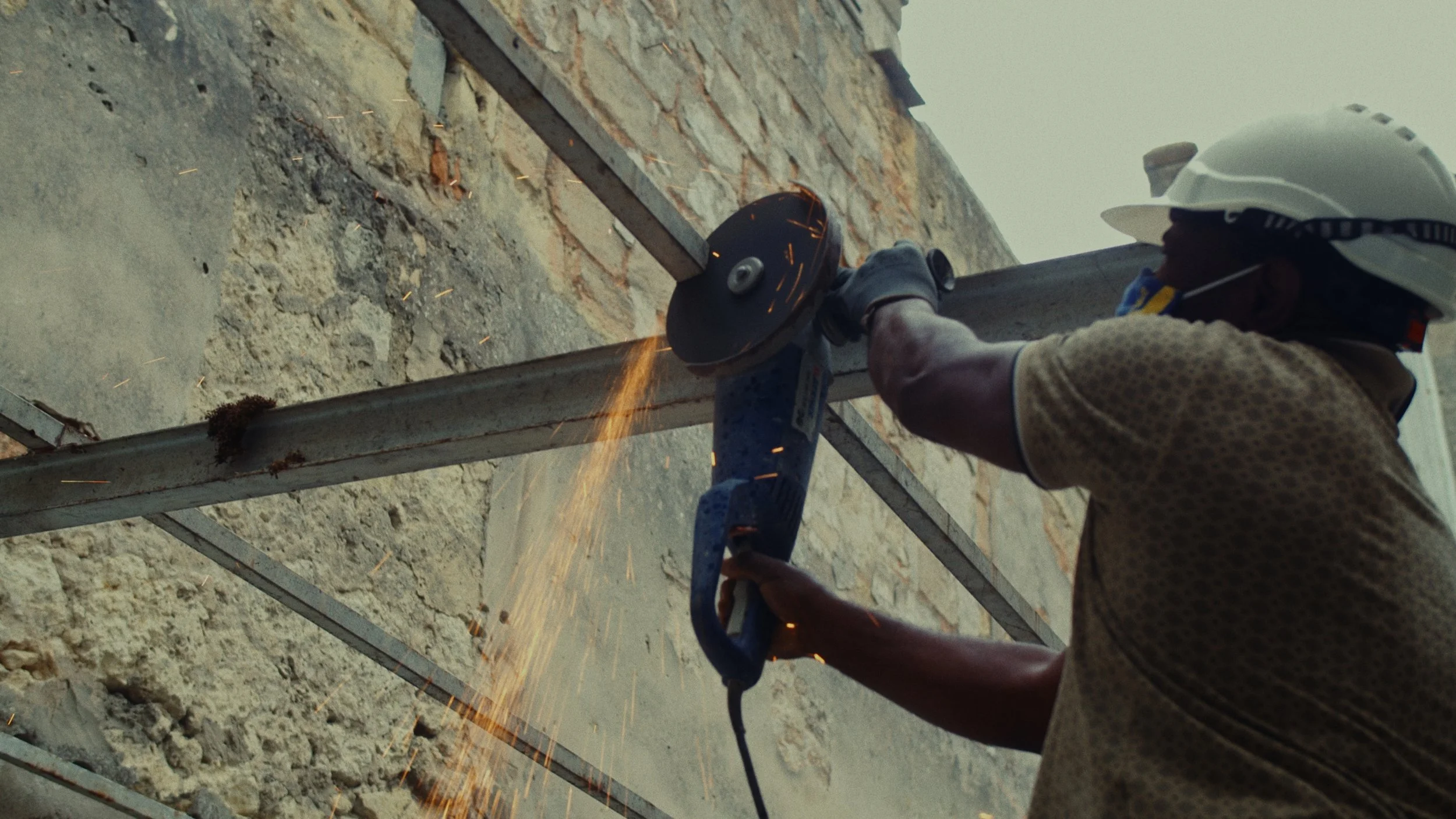 Un ouvrier portant un casque de sécurité utilise une disqueuse pour couper une pièce de métal, avec des étincelles qui volent.