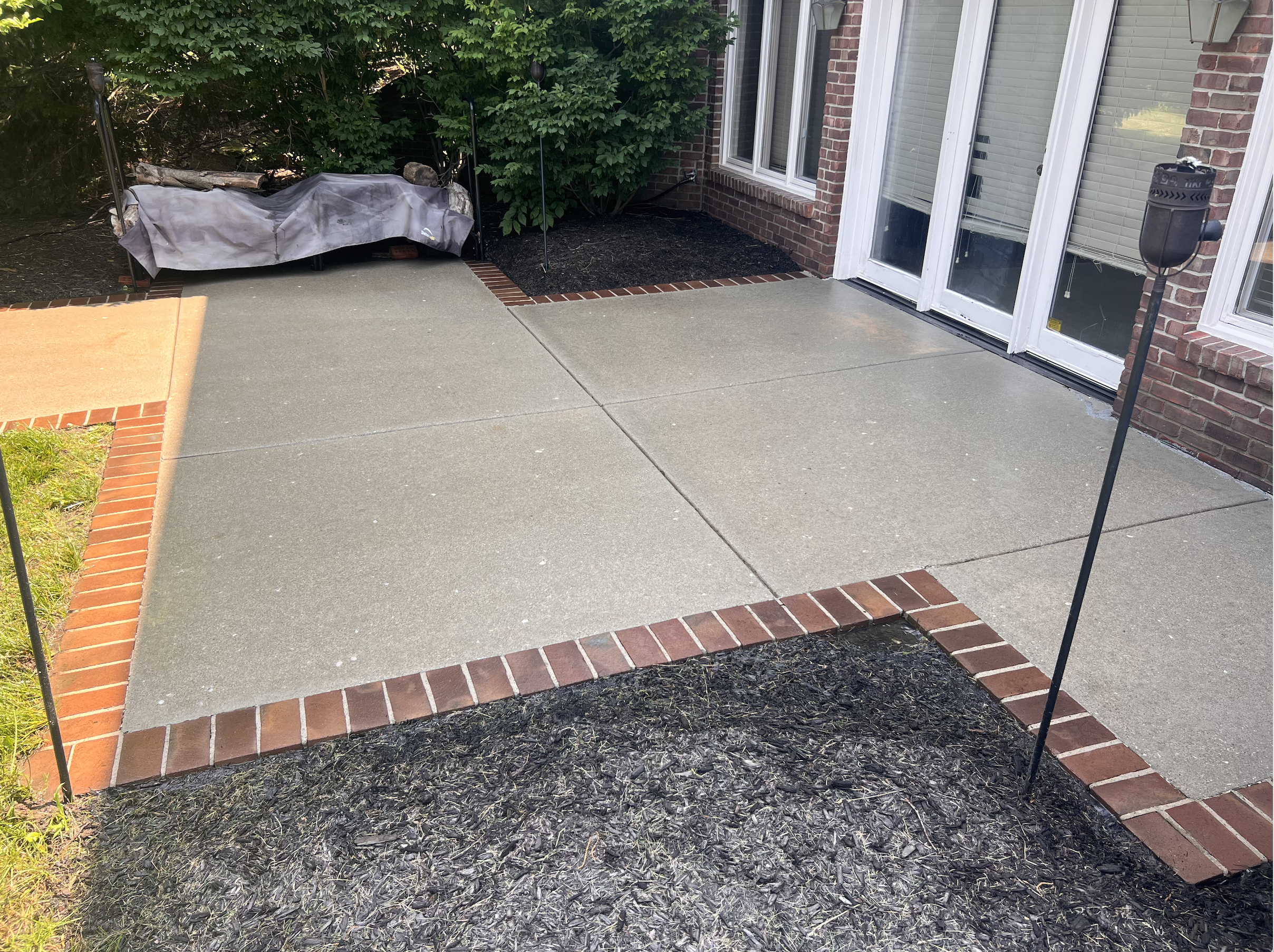 Newly poured concrete patio with brick border next to a brick house with sliding glass door, black lanterns, and a covered outdoor furniture piece, with greenery in the background.