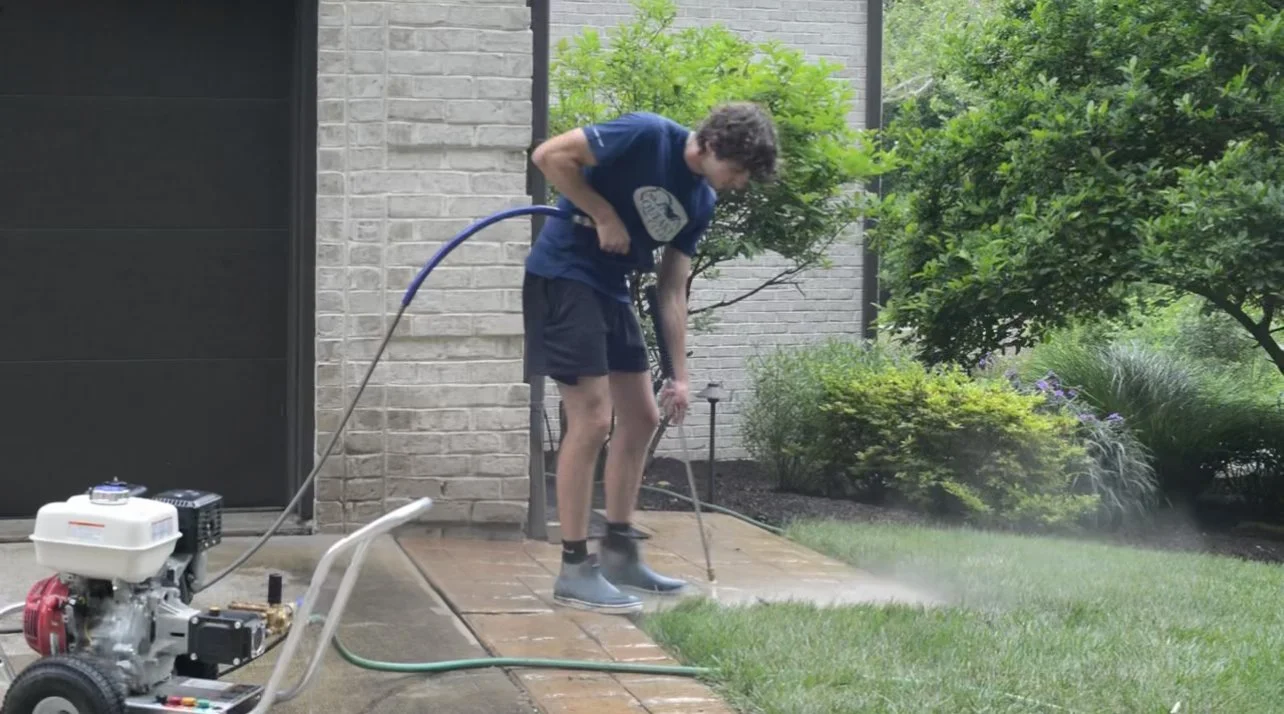 A young person wearing a blue t-shirt, shorts, and boots, standing on a brick patio and using a pressure washer to clean the grass and pavement outdoors.