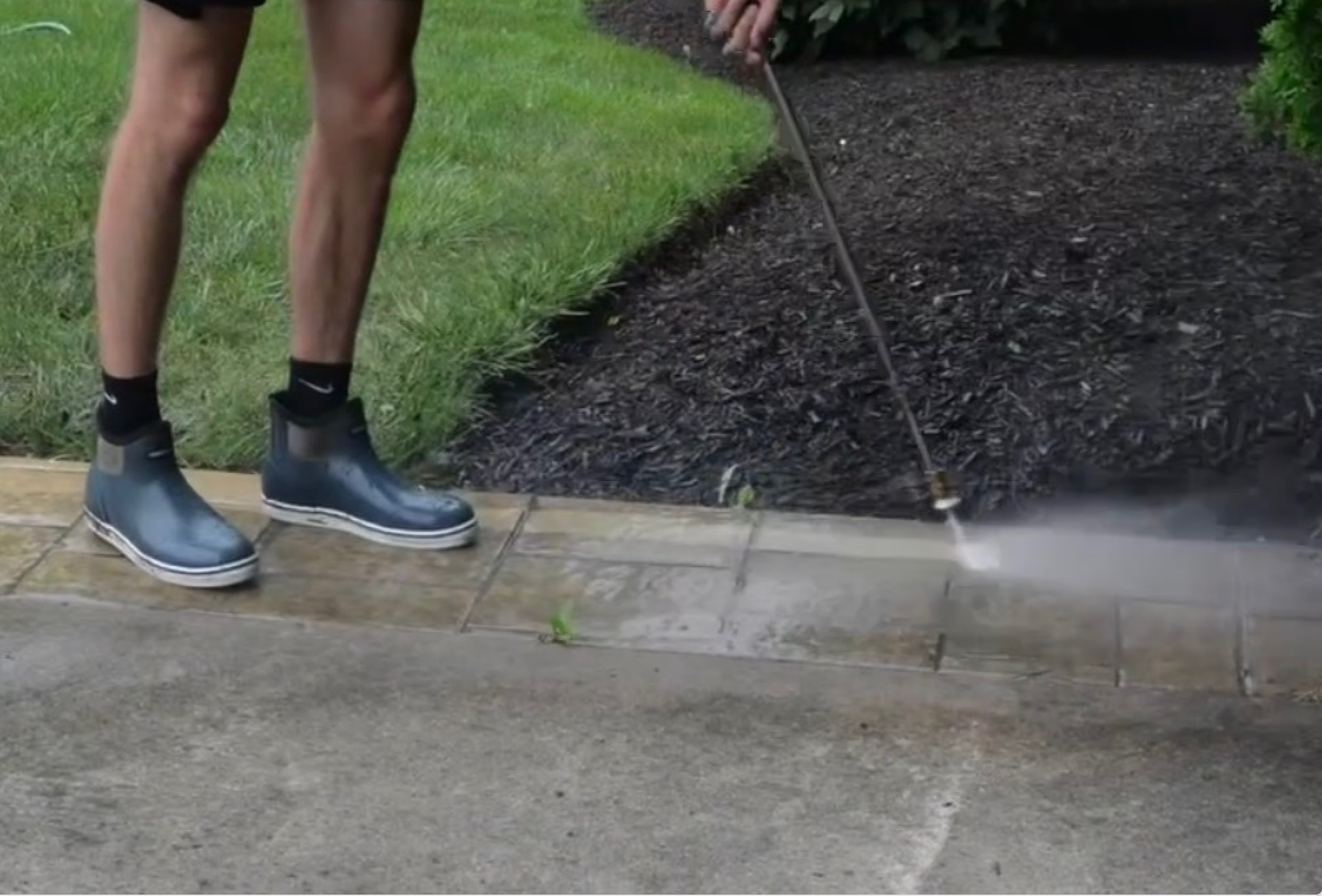Person using a garden sprayer to spray a weed killer on a garden bed with mulch and grass nearby.
