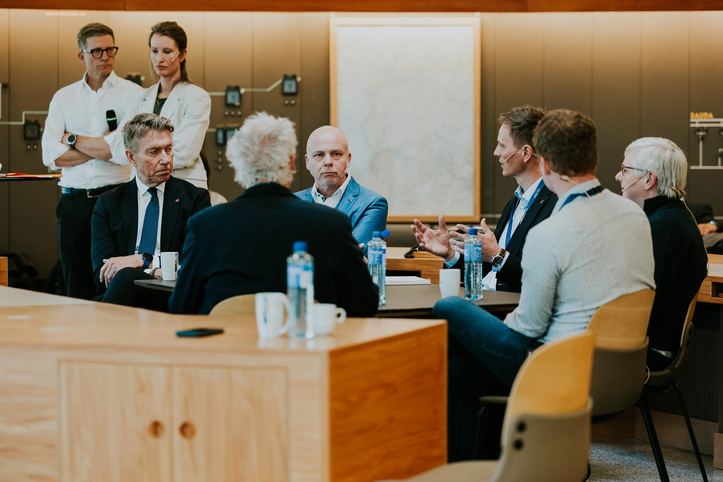 Et møte med syv personer, noen med mikrofon, i et konferanserom. Flere vannflasker og kopper på bordet.