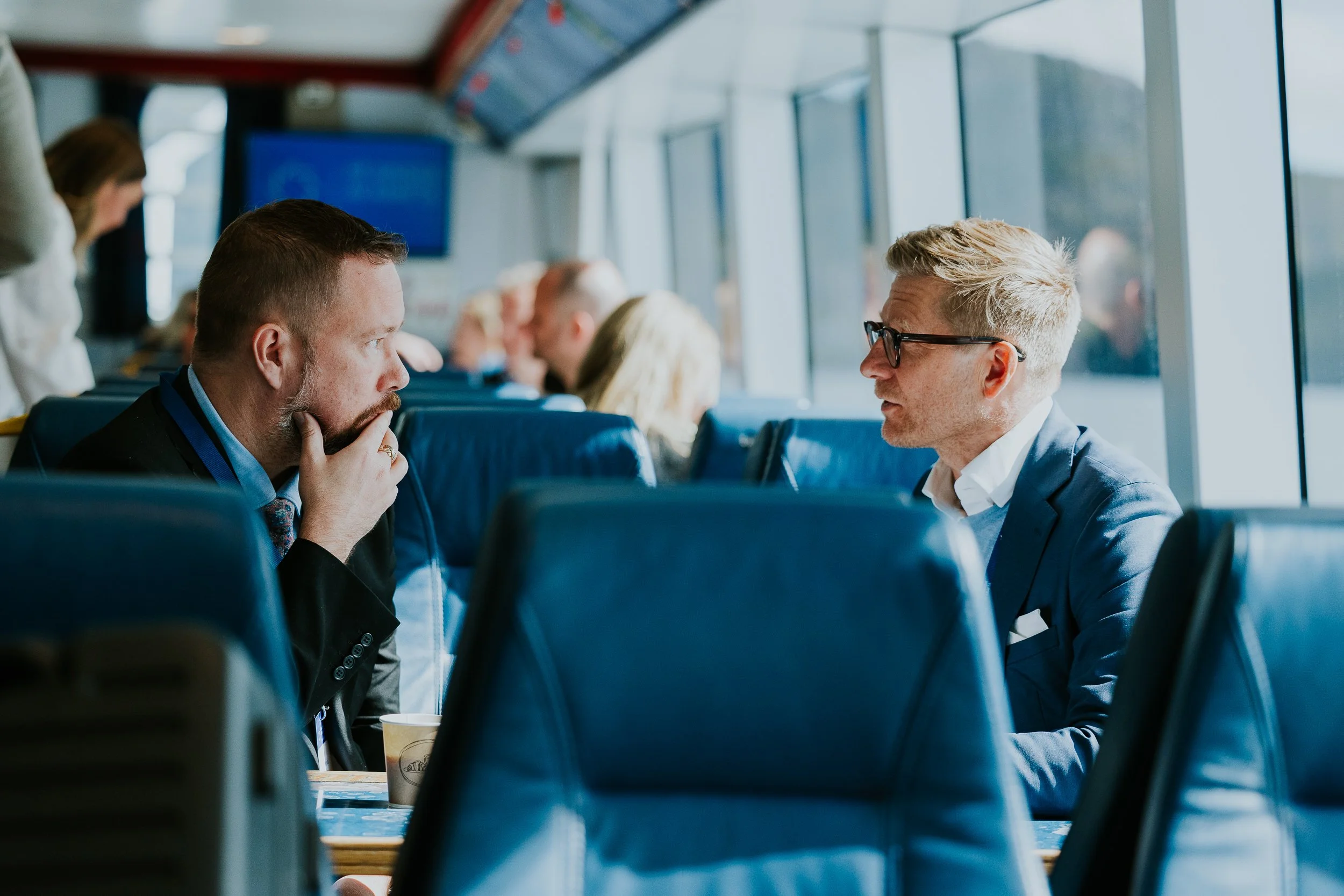 To menn sitter mot hverandre på et flysete. Den ene mannen har skjegg, har på seg en svart dress og shirt, og ser tankefull ut. Den andre mannen har blond, kort hår, bruker briller og en blå dress.